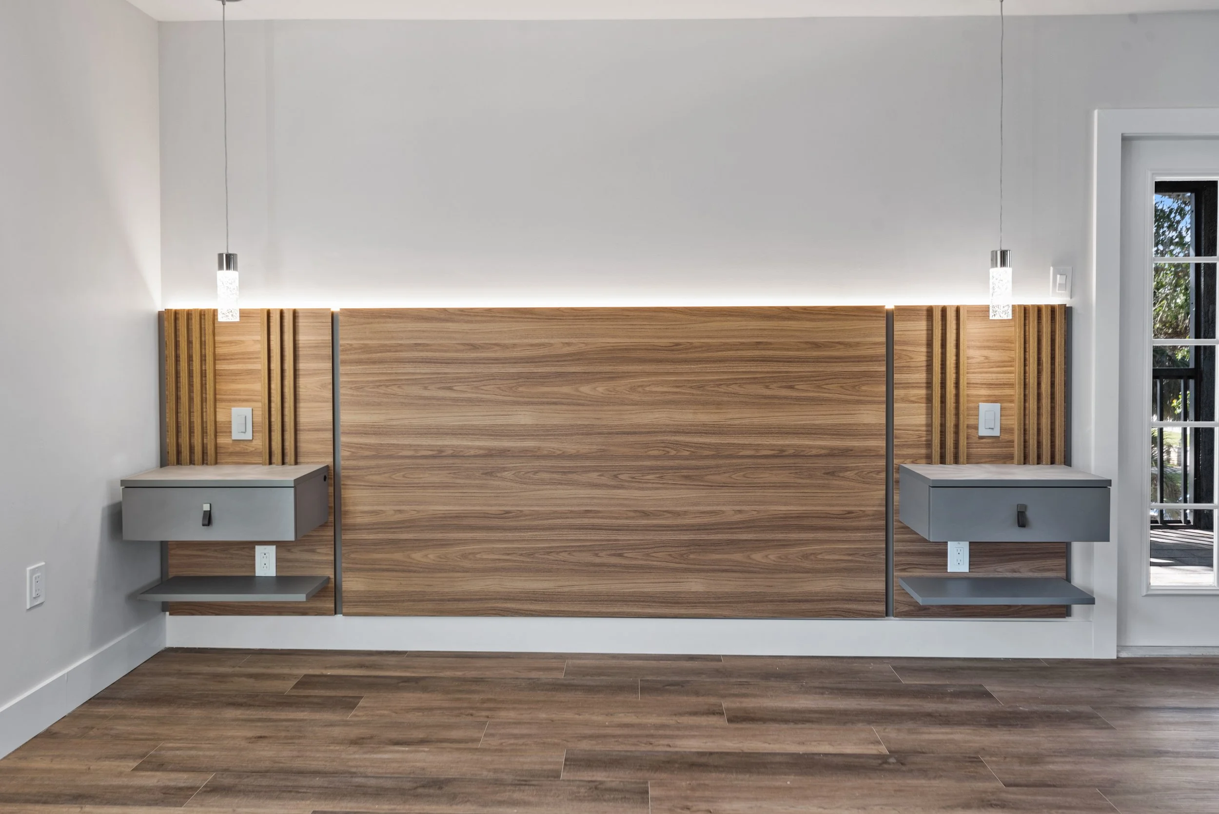 Modern bedroom headboard with wooden panel, two nightstands, pendant lights, and a door with a window to the right.