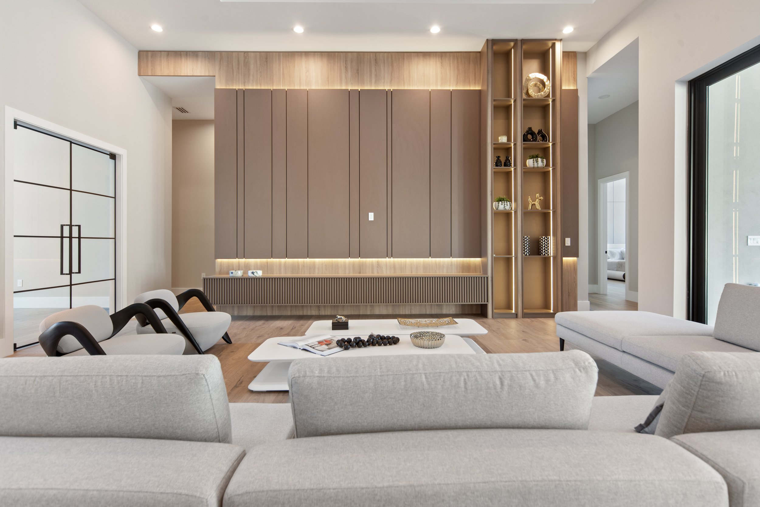 Modern living room with a beige sectional sofa, two white armchairs with black accents, a white coffee table with decorative items, a built-in wall unit with brown panels and open shelves, and sliding glass doors leading outside.