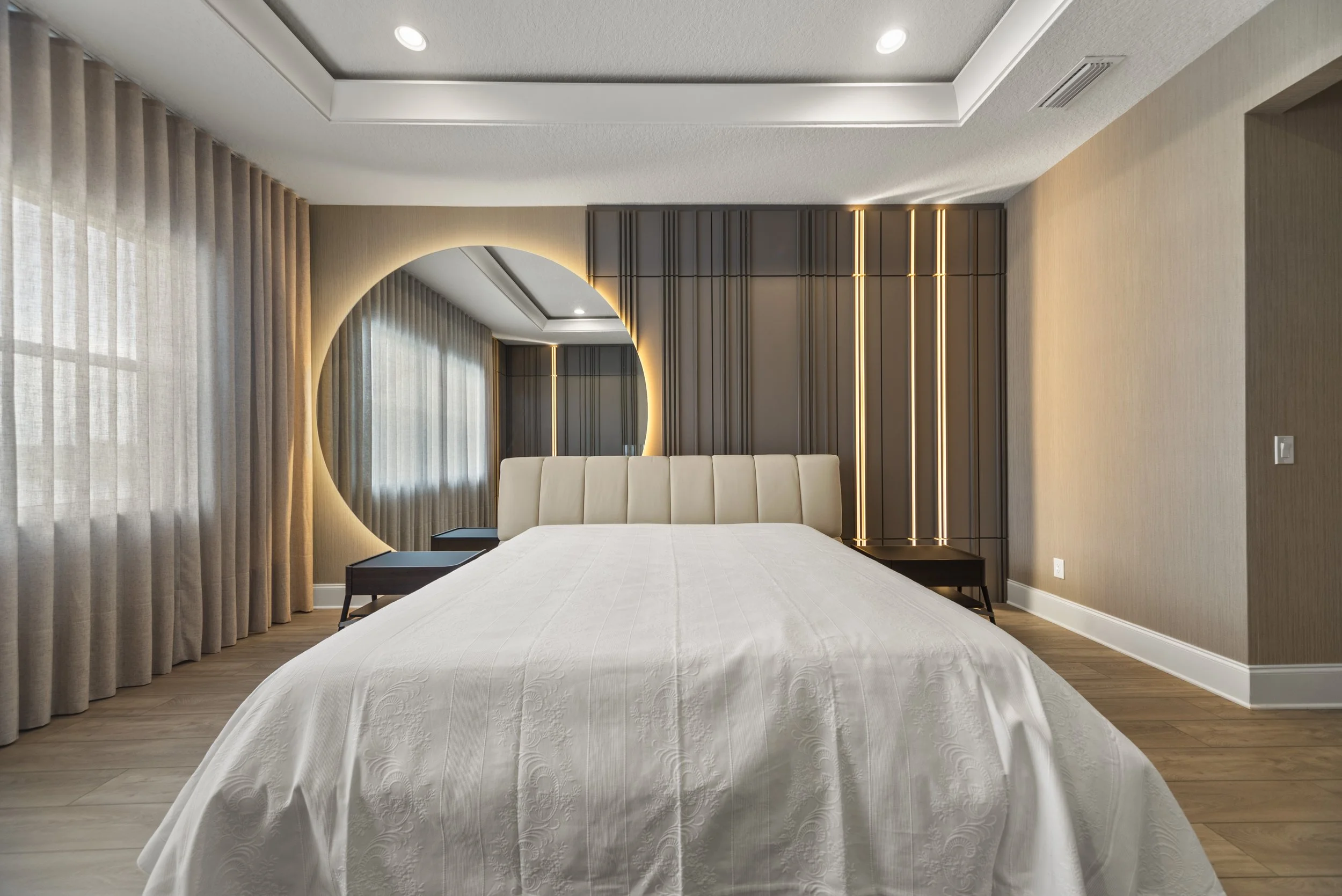 Modern bedroom with a large bed featuring a cream headboard, a round mirror on the wall with a soft backlight, dark wooden bedside tables, beige curtains, and warm lighting accents on a dark gray wall panel.