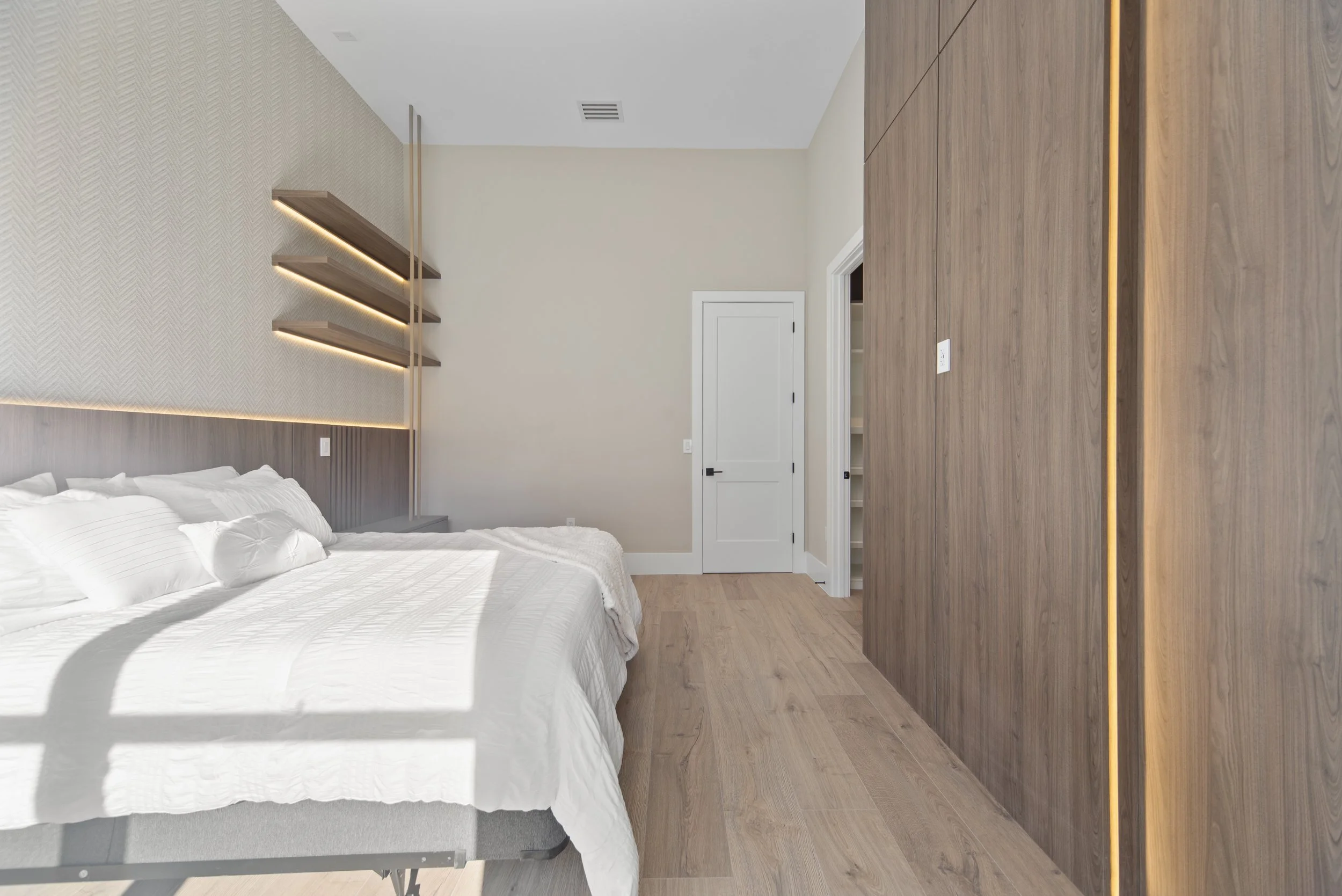 Modern bedroom with a large bed dressed in white linens, a wood-paneled wall with subtle lighting, and an open closet with shelves.
