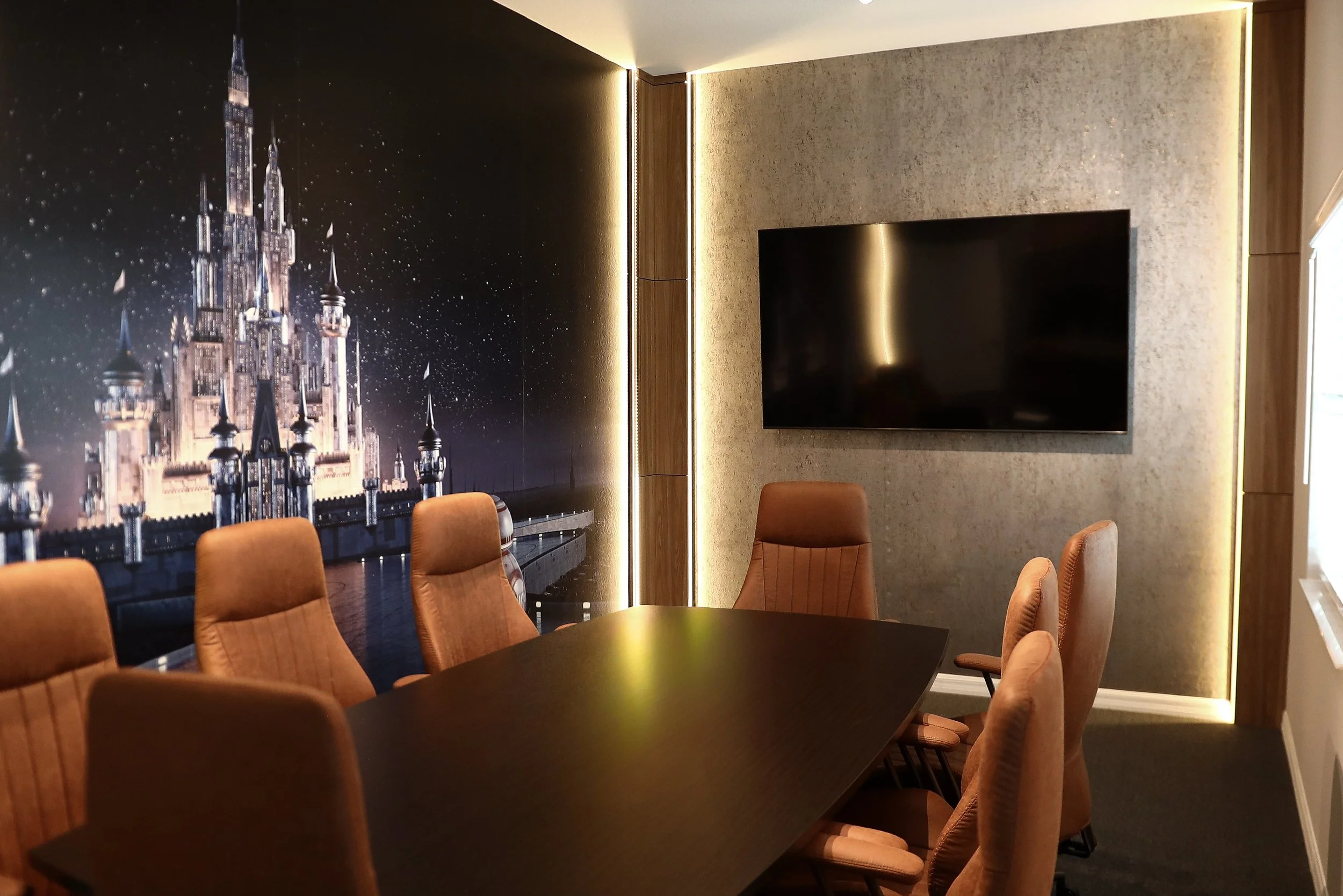 A modern conference room with a black table, tan padded chairs, a wall-mounted television, a decorative wall with a castle mural, and warm lighting.
