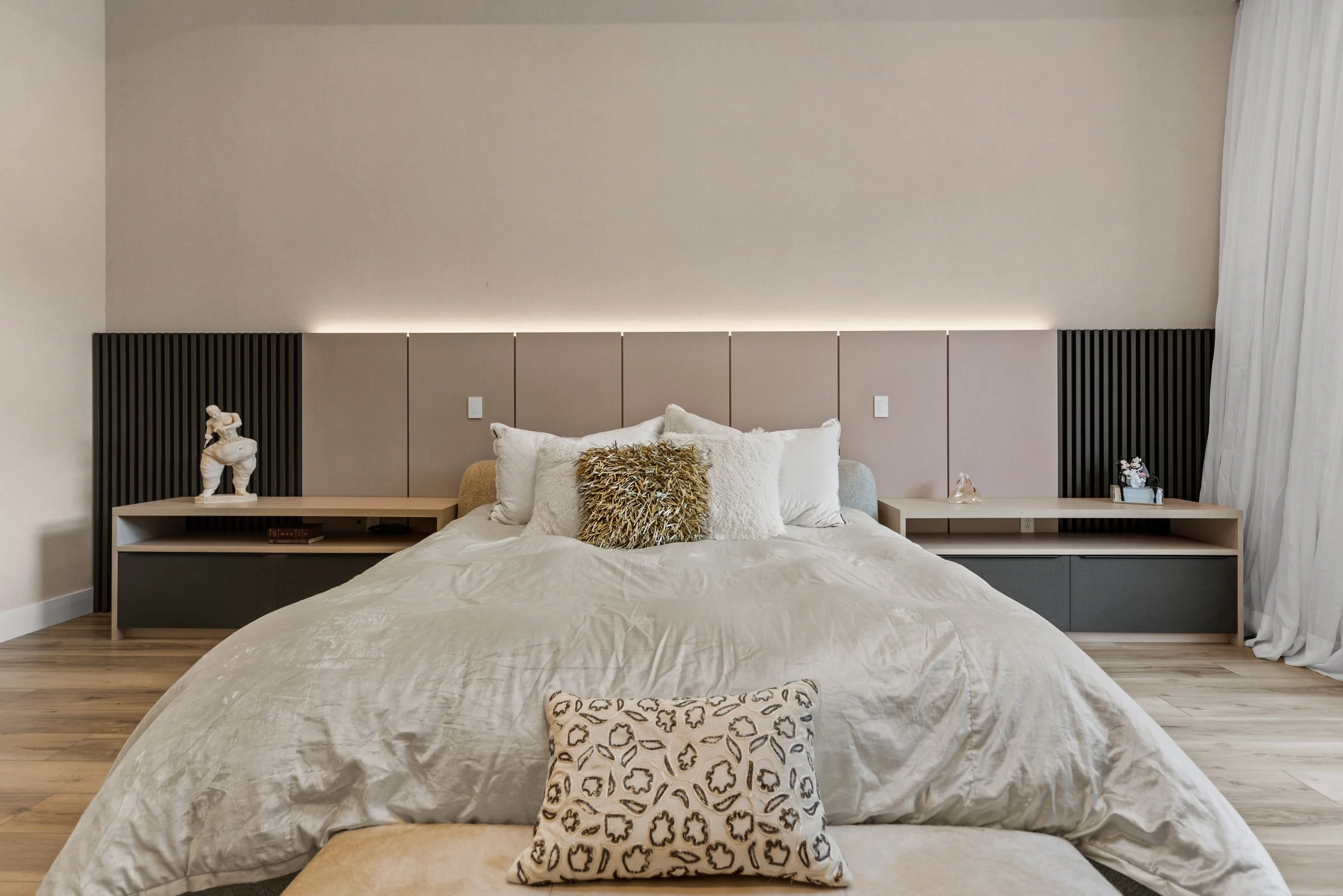 Modern bedroom with a large bed, beige upholstered headboard, and neutral-toned bedding. Decorative pillows include a fuzzy white pillow and a patterned leopard print pillow. Two minimalist nightstands on each side hold decor items, with a sculpture 
