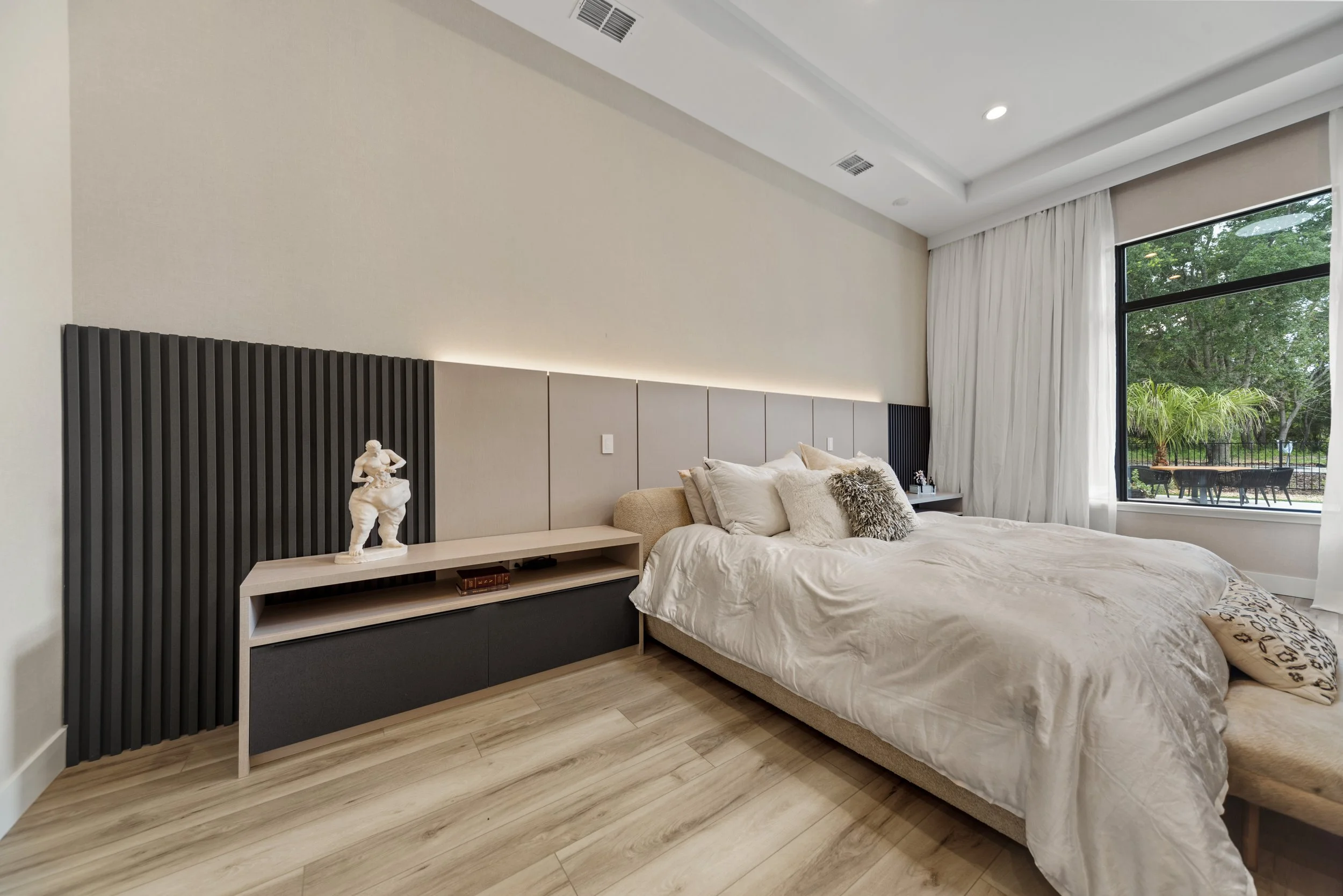 Modern bedroom with a large bed, white bedding, and decorative pillows. There is a view of a backyard with outdoor furniture through a large window.