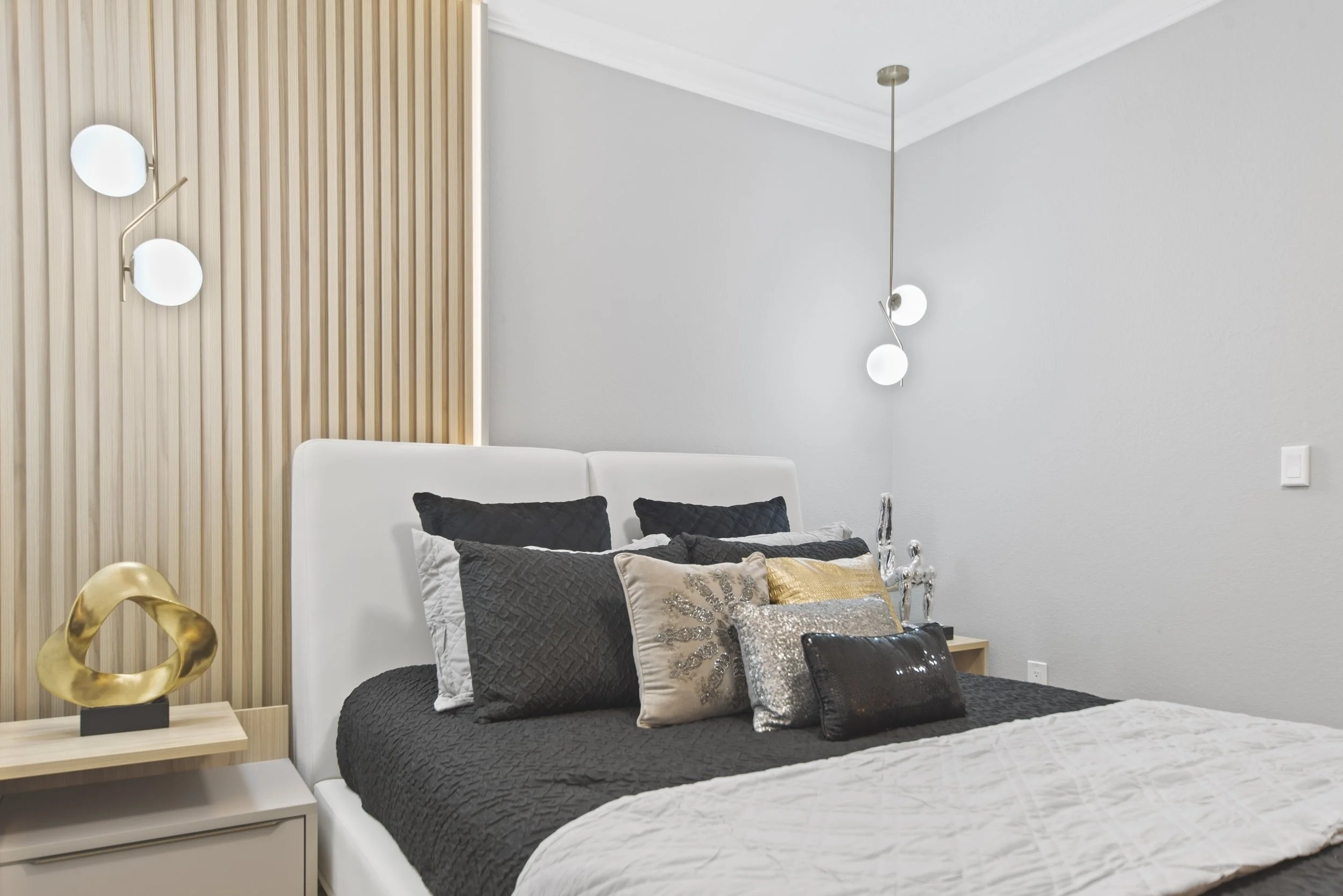 Modern bedroom with a white bed, decorative pillows, wooden side table with a gold abstract sculpture, contemporary wall lights, and neutral wall color.