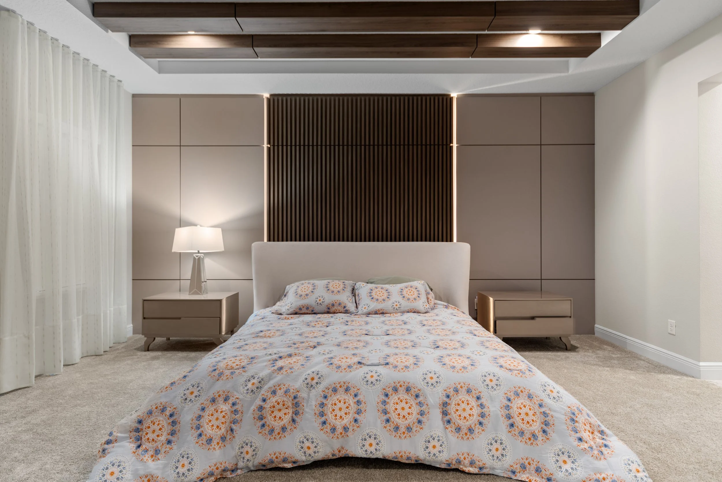 Modern bedroom with beige walls, a bed with patterned duvet, two nightstands, a table lamp, and decorative wooden panels on the ceiling and wall.