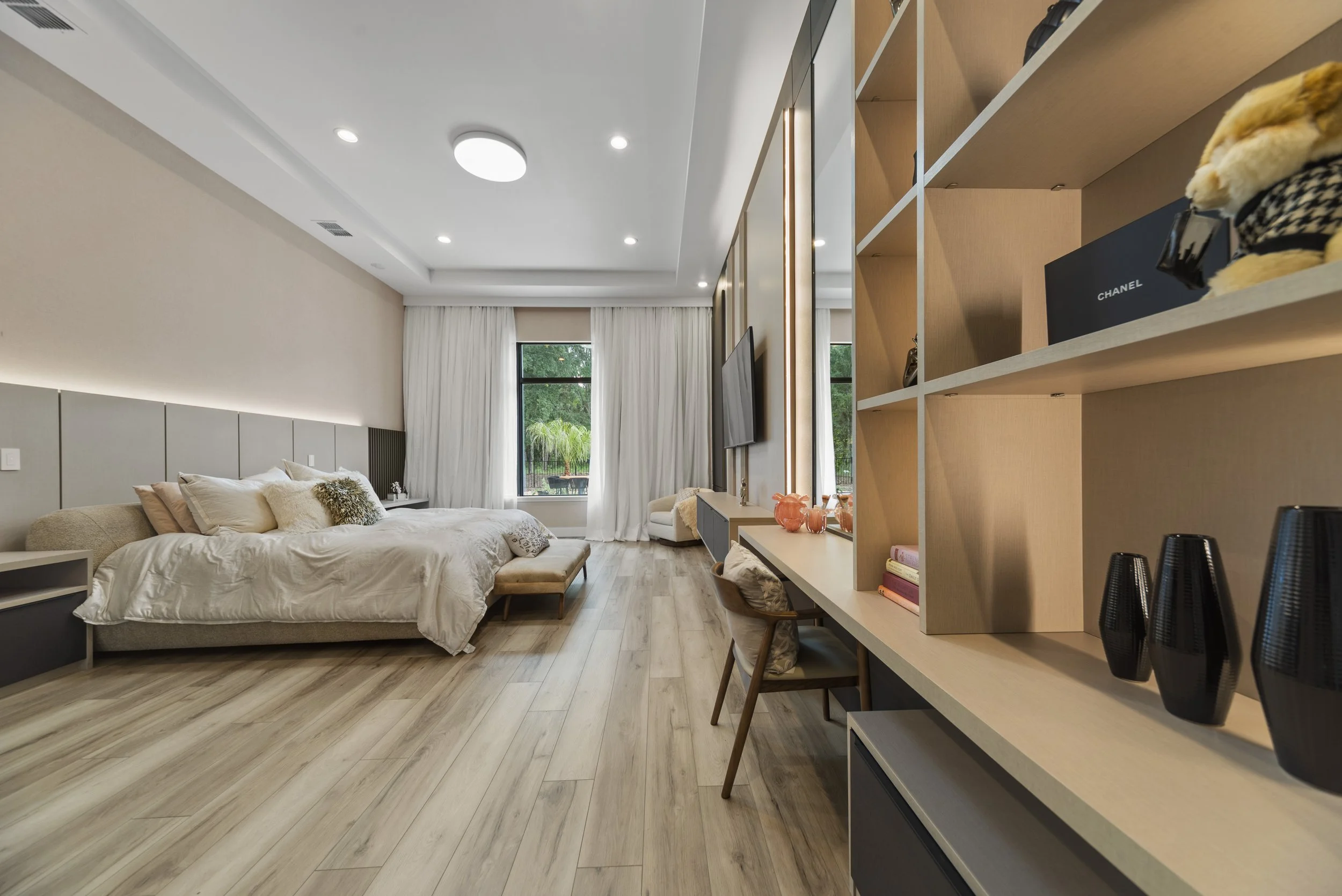 Modern bedroom with a large bed, white bedding, and pillows, a window with white curtains, a wall-mounted TV, a beige chair, and a built-in bookshelf with decorative items, books, and a Chanel box.