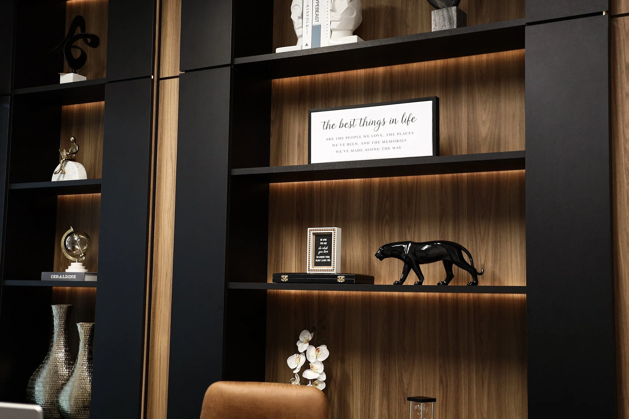Decorative bookshelf with black and wooden shelves, featuring a framed quote, animal figurines, vases, and a flower arrangement.