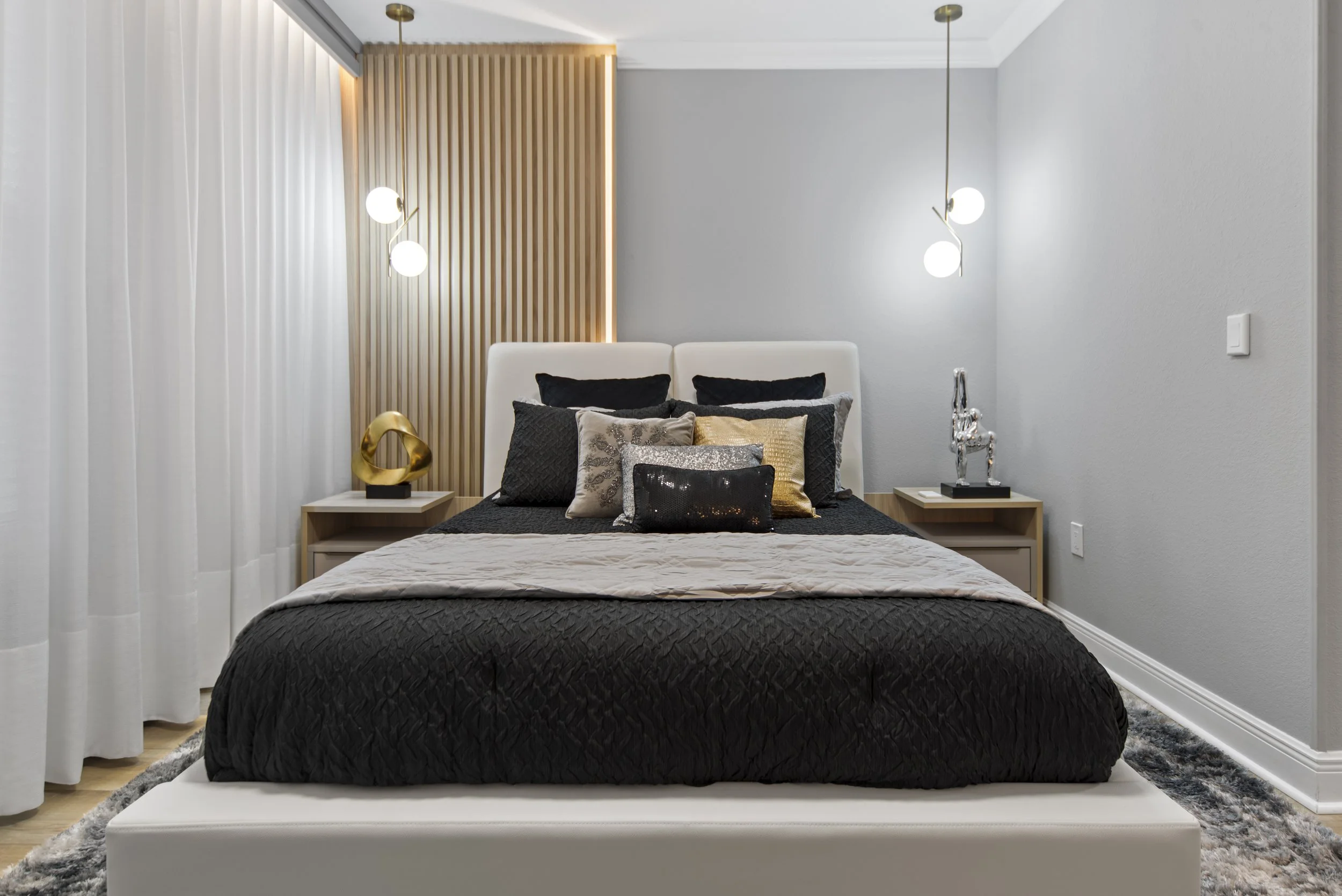 Modern bedroom with a white bed frame, black and gray bedding, decorative pillows, two nightstands with sculptures, and wall-mounted lights.