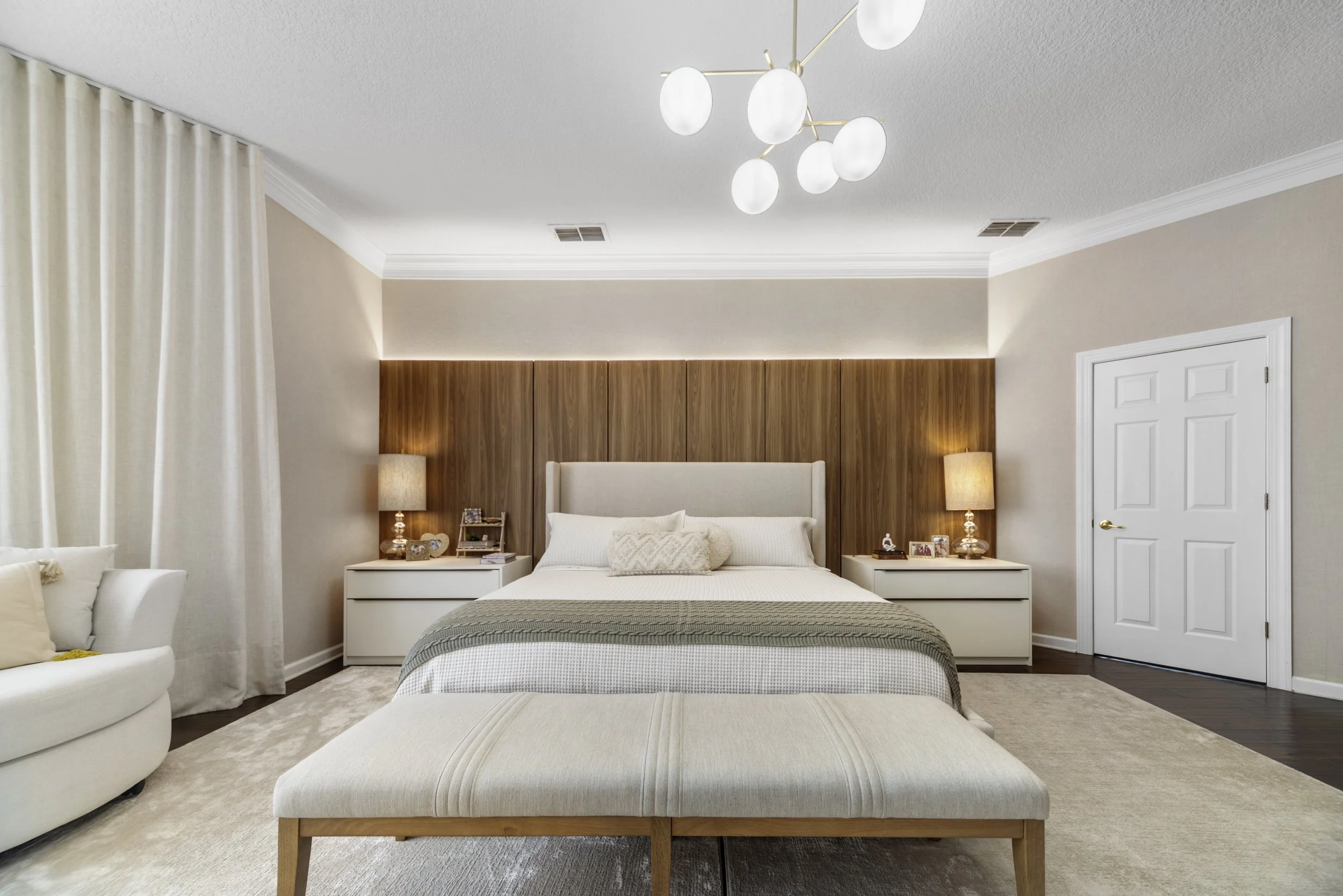 A modern bedroom with a large bed, beige walls, wooden headboard, nightstands with lamps, a bench at the foot of the bed, a white armchair, and a ceiling light fixture.