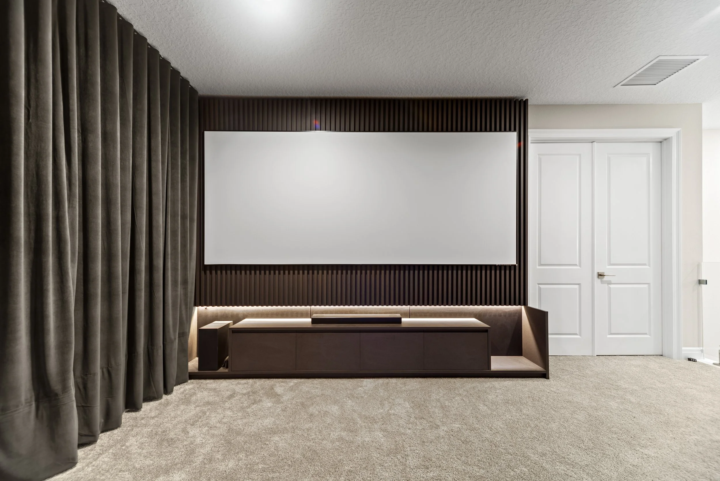 Modern living room with a large blank white television screen on a black textured wall, a dark wooden media console underneath, beige carpet, and white double doors to the right.