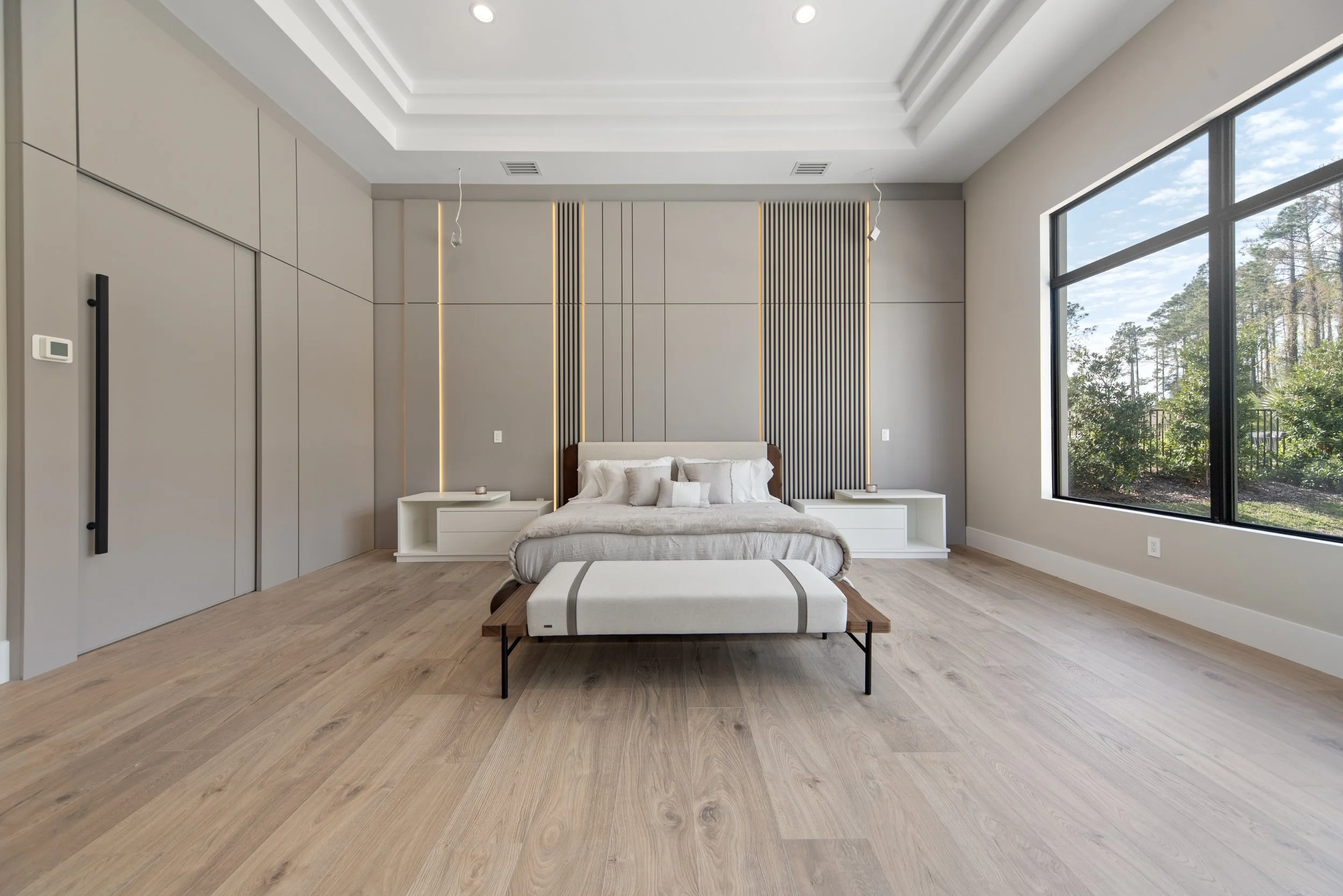 Modern bedroom with light wood flooring, a bed with a beige headboard, white bedding, and an ottoman at the foot of the bed. Large window on the right with a view of trees outside. Minimalist nightstands on each side of the bed, with a small item on 