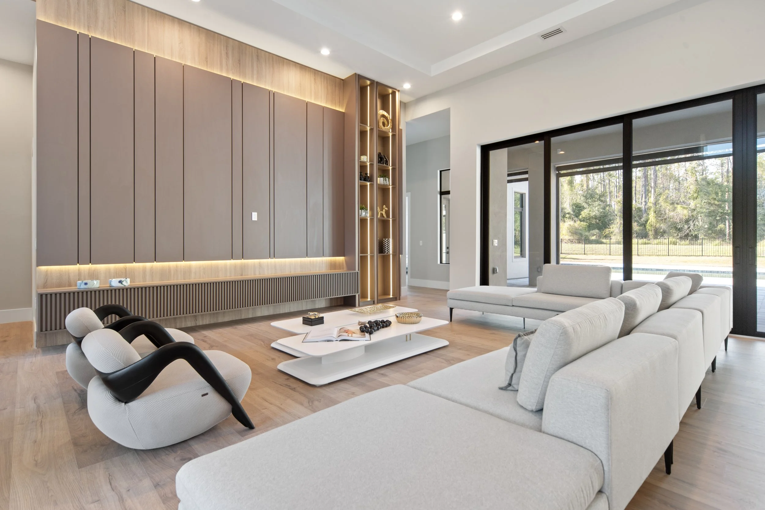 Modern living room with beige sectional sofa, white coffee table, black and white lounge chairs, large windows, and a wooden accent wall with built-in shelving.