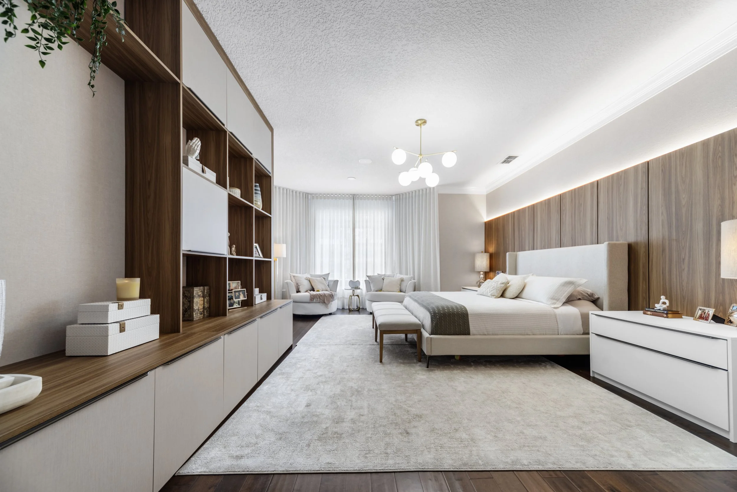 Modern bedroom with a large bed, white headboard, white dresser, wooden accent wall, white curtains, sitting area with armchairs, and a wooden shelf unit.