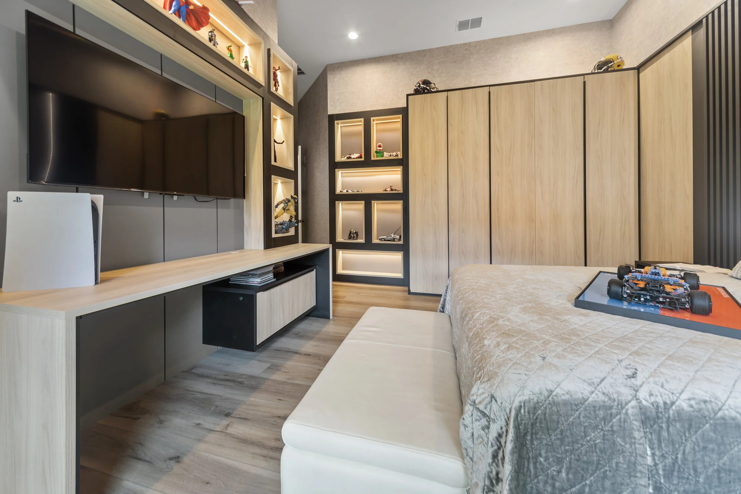 Modern bedroom with wooden wardrobe, display shelves with LED lighting, wall-mounted flat-screen TV, and a toy race car on the bed.