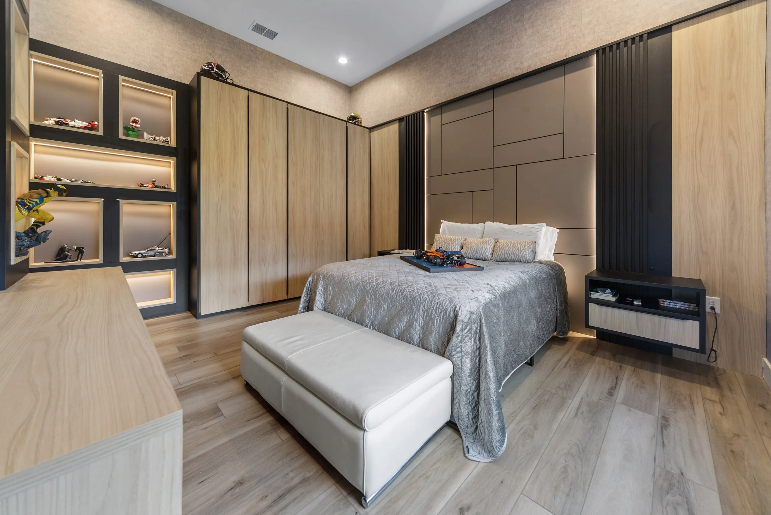 Modern bedroom with a bed covered in a silver quilt, a white ottoman at the foot, built-in wooden wardrobes, a black and wood TV stand, and a display shelf with toy cars and models.