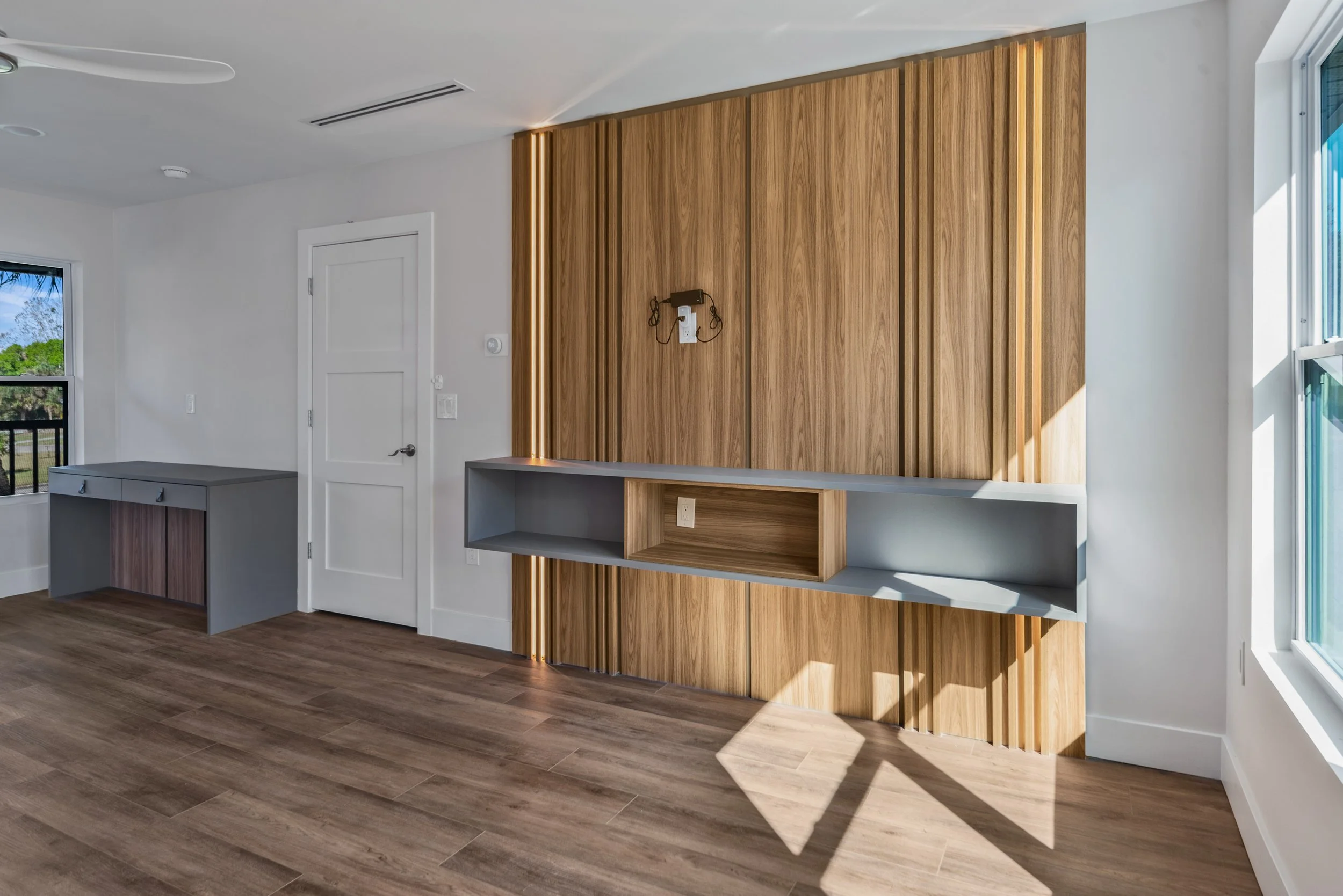 Minimalist living room with a large wooden accent wall with integrated lighting, gray built-in cabinets, and a white door. Sunlight casts shadows through a window on wooden flooring.