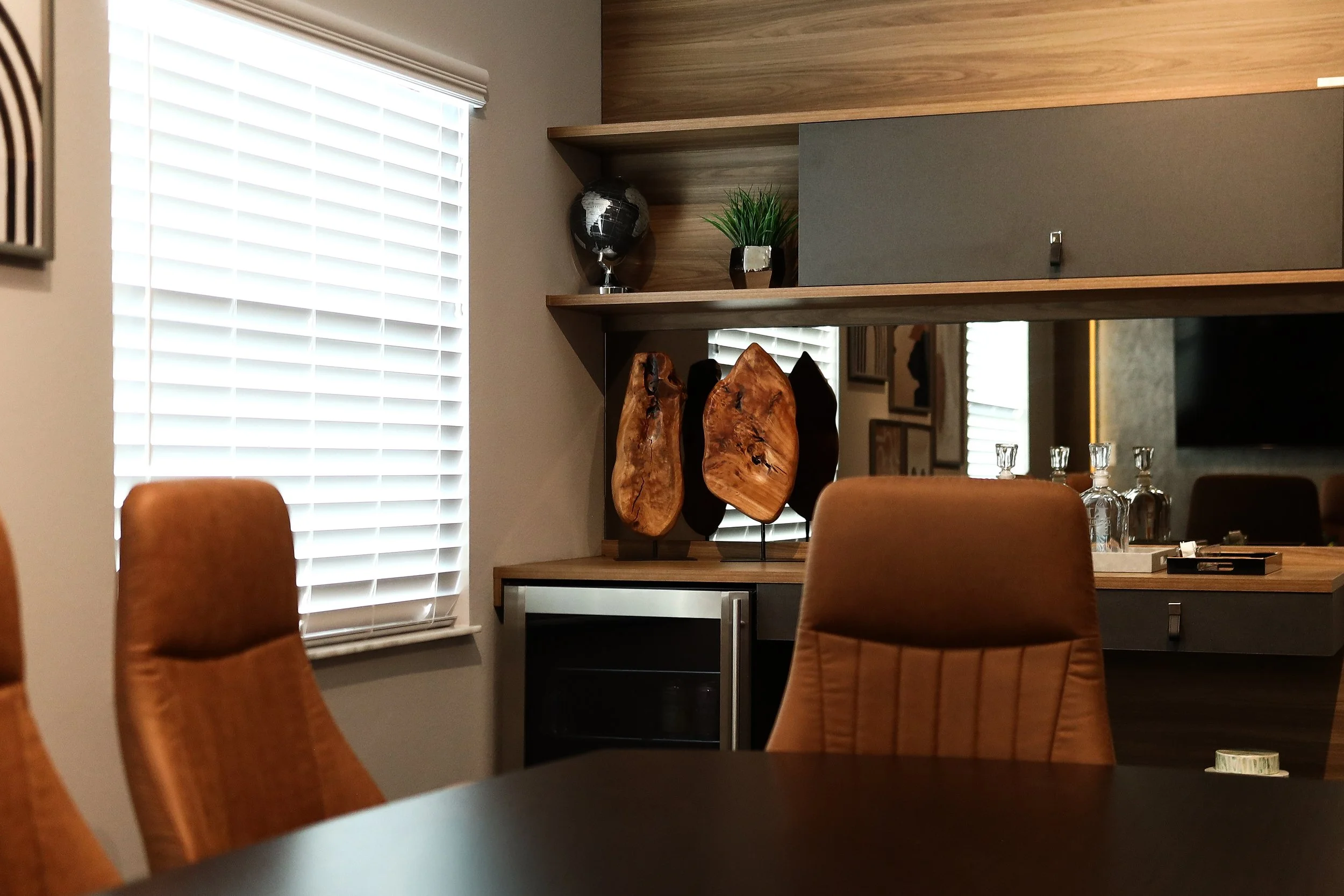 A modern dining room area with a black table, brown leather chairs, window with white blinds, a built-in wooden shelf with decorative items, and a mirror reflecting part of the room.