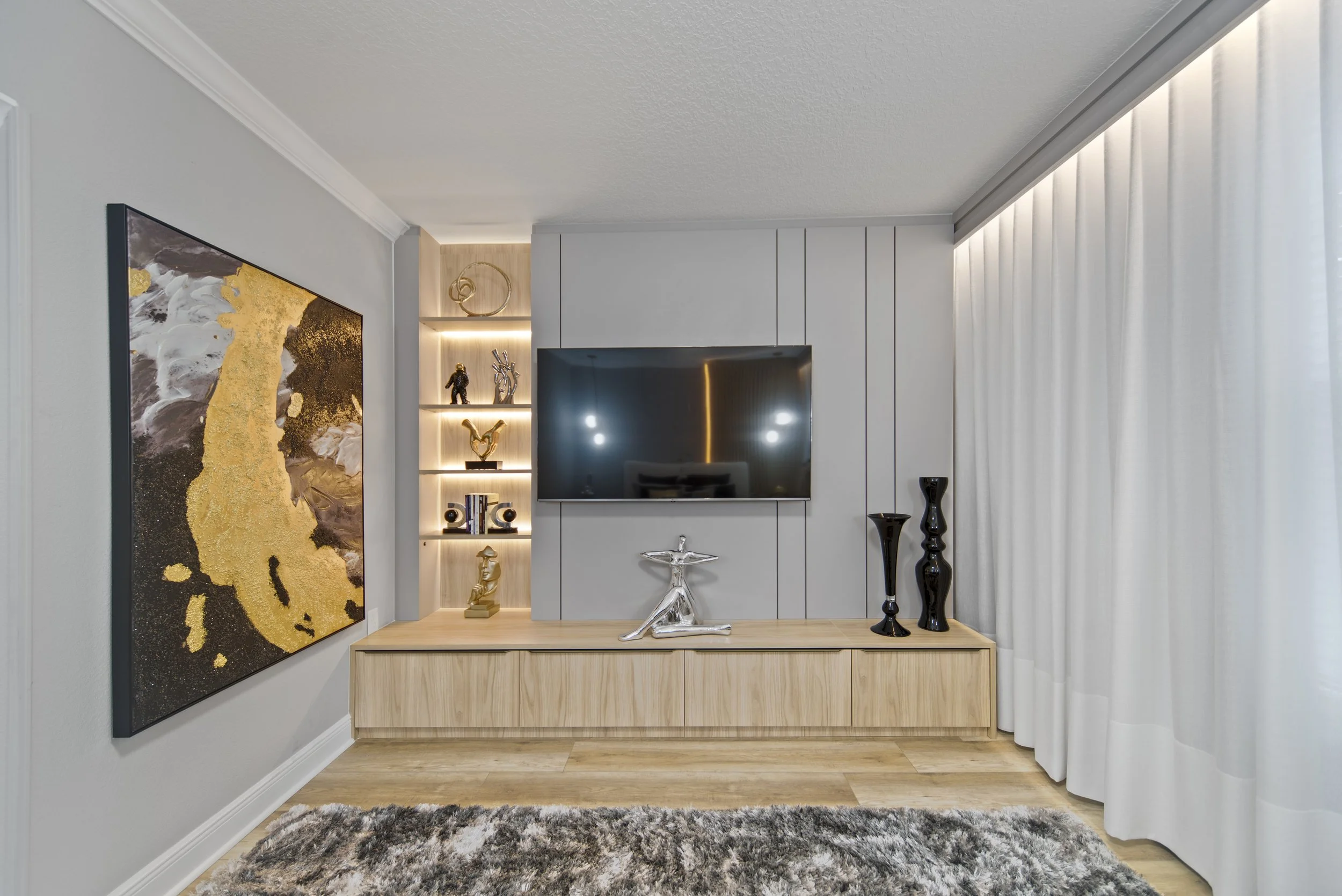 Modern living room with wall-mounted flat-screen TV, decorative sculptures on a wooden cabinet, abstract artwork on the wall, built-in shelving with decorative items, and cream-colored curtains.