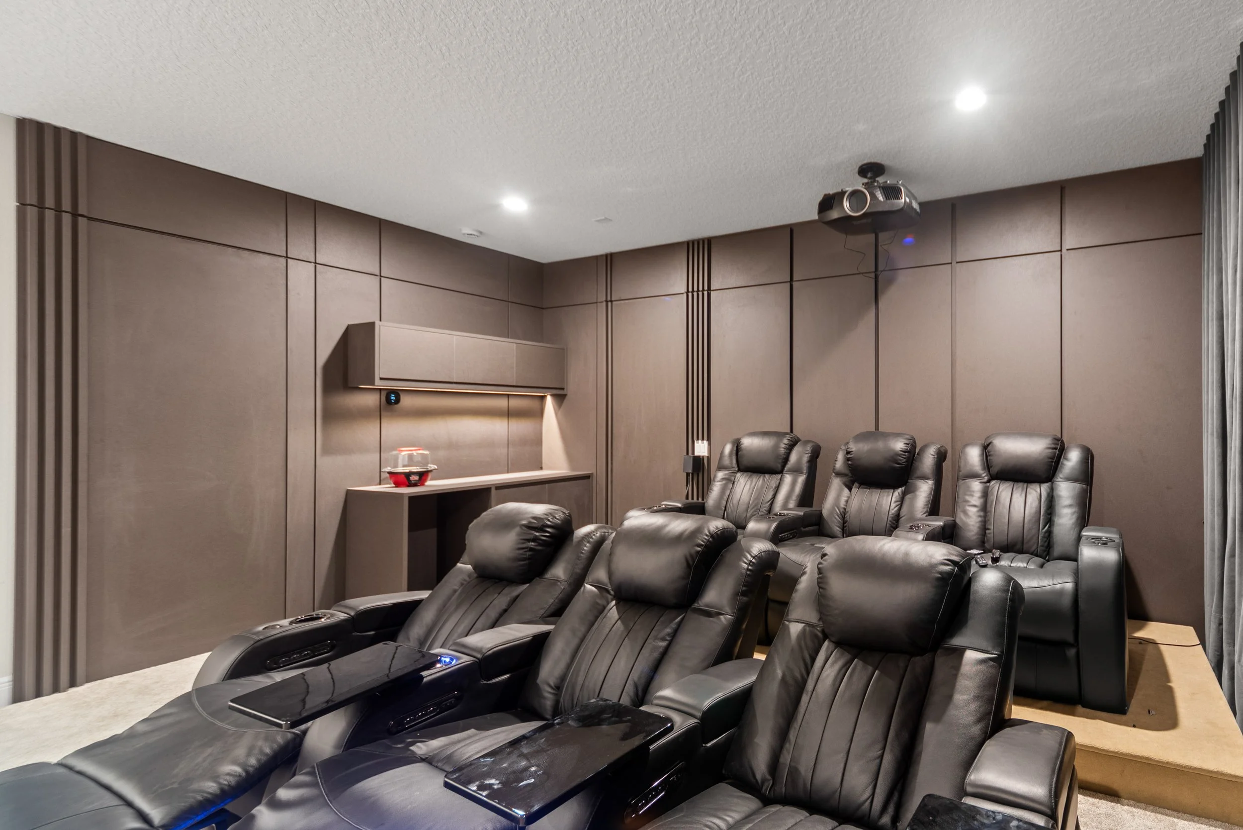 A modern home theater room with six black leather reclining chairs, a projector mounted on the ceiling, and brown wall panels.
