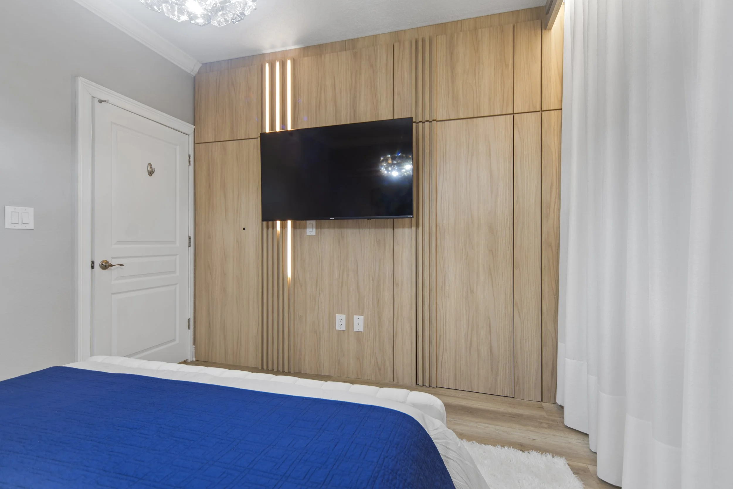 A bedroom with a wall-mounted TV on a wooden panel wall, a bed with white and blue linens, a closed white door, and white curtains on the right side.