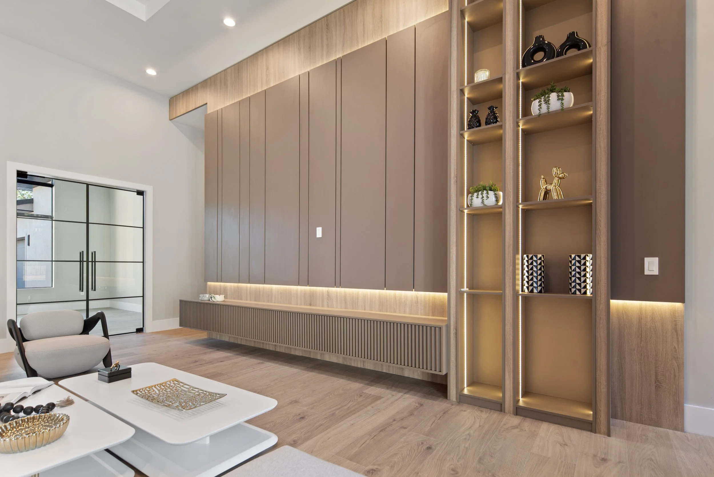 Modern living room featuring a built-in wood panel wall with concealed lighting, a glass sliding door, a white table with decorative items, and a wooden shelving unit with decorative sculptures and plants.