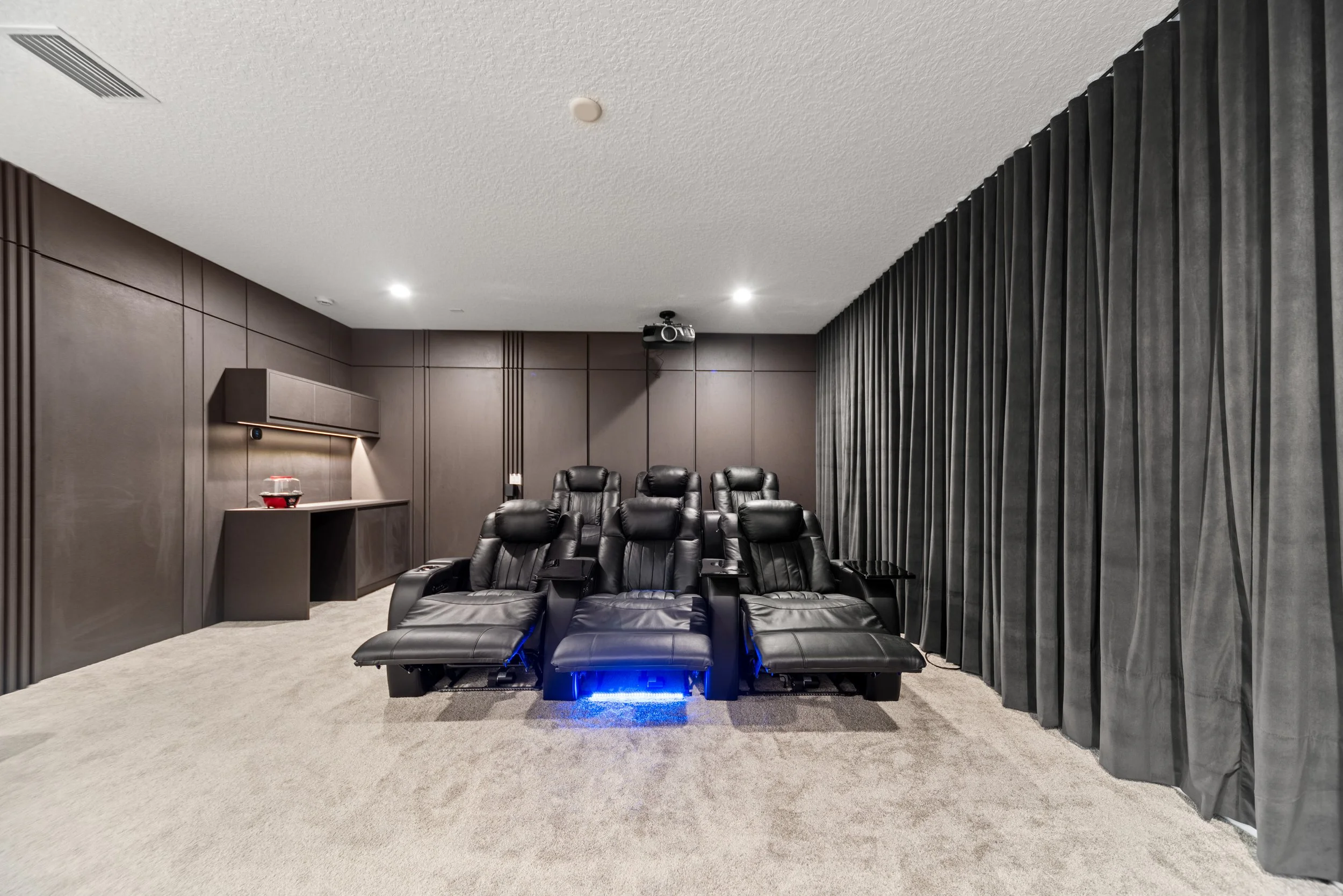 Home theater room with black leather recliner seats, dark wood panel walls, and a projection screen. There is a side table with a bowl on it and black curtains on one side.