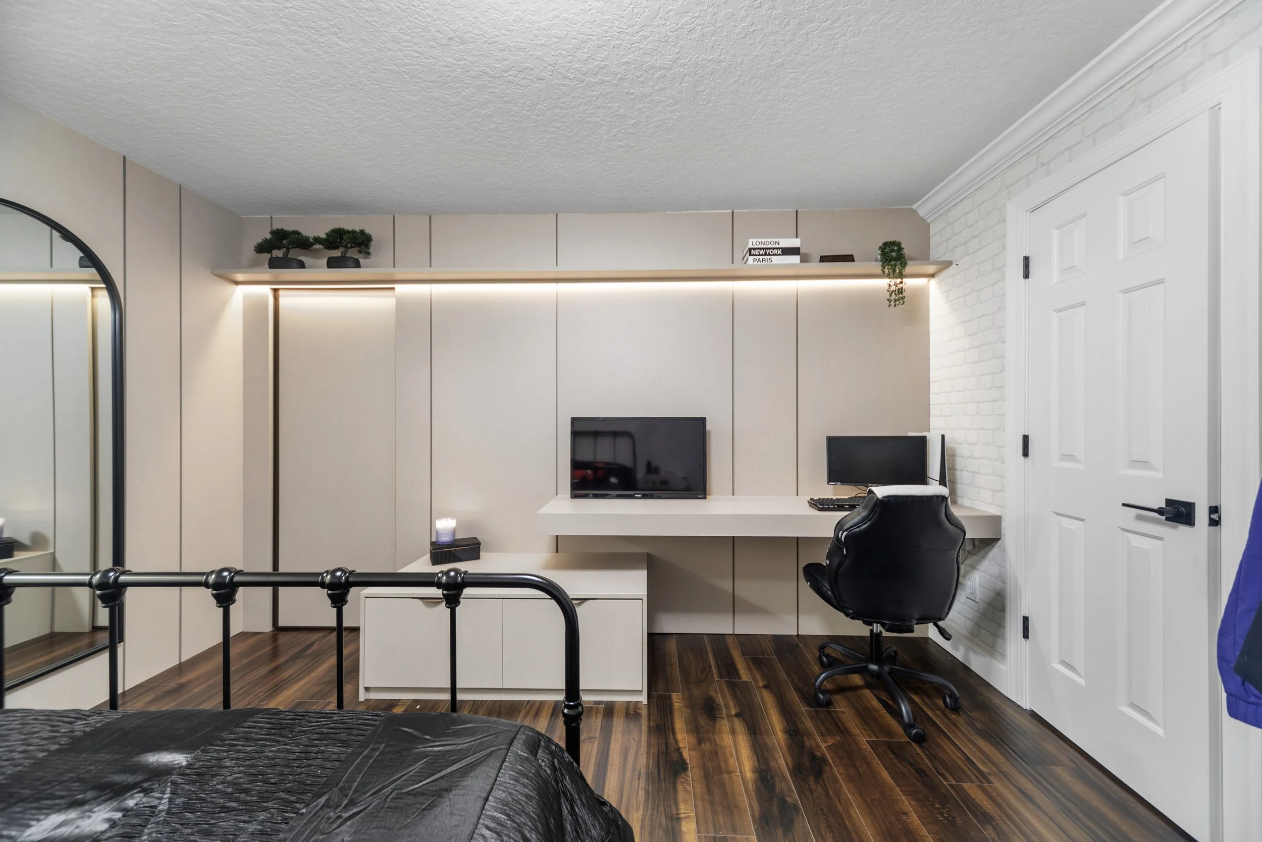 Modern bedroom workspace with a white desk, two monitors, a black swivel chair, and decorative plants on a shelf. Part of a bed with a black blanket is visible in the foreground. The room has wooden flooring and white walls with a brick accent.