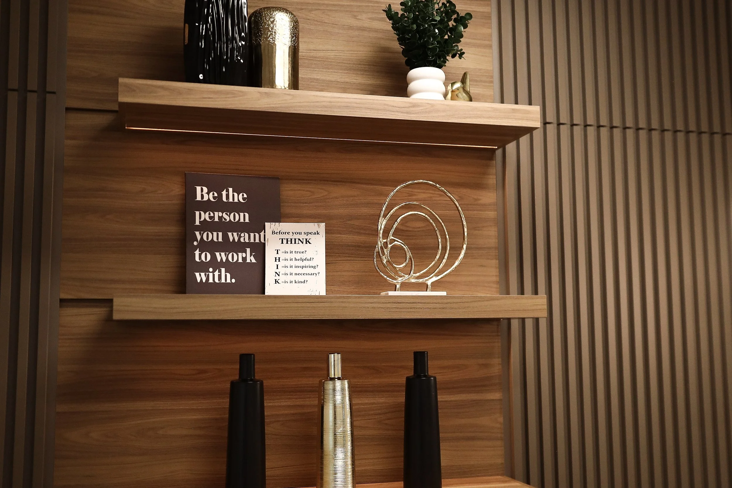 Decorative display on wooden shelves with black, white, and gold vases, a small green plant in a white pot, and inspirational signs. The signs include one black sign with white text that says, 'Be the person you want to work with,' and a white sign w
