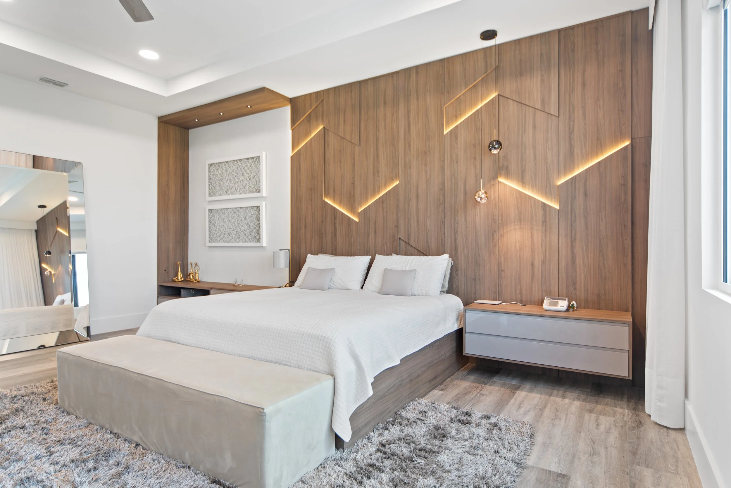 Modern bedroom with a large bed with white bedding and two pillows, a wooden accent wall with geometric light fixtures, a window with white curtains, a mirrored dresser, a nightstand with a phone and book, a shaggy area rug, and minimal decor.
