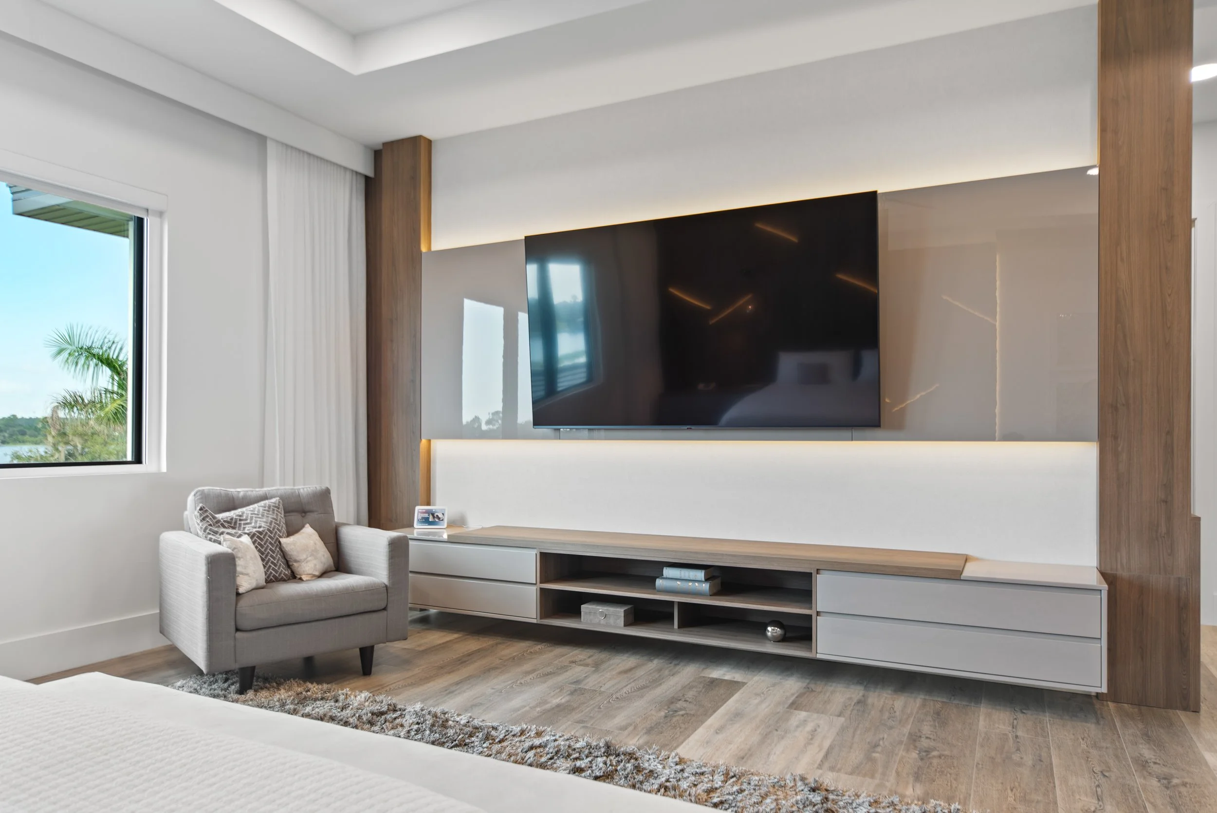 Modern living room with a large wall-mounted TV, a grey armchair with decorative pillows, and a sleek white and wood media console. There is a window with a view of palm trees outside and neutral-toned wooden flooring.