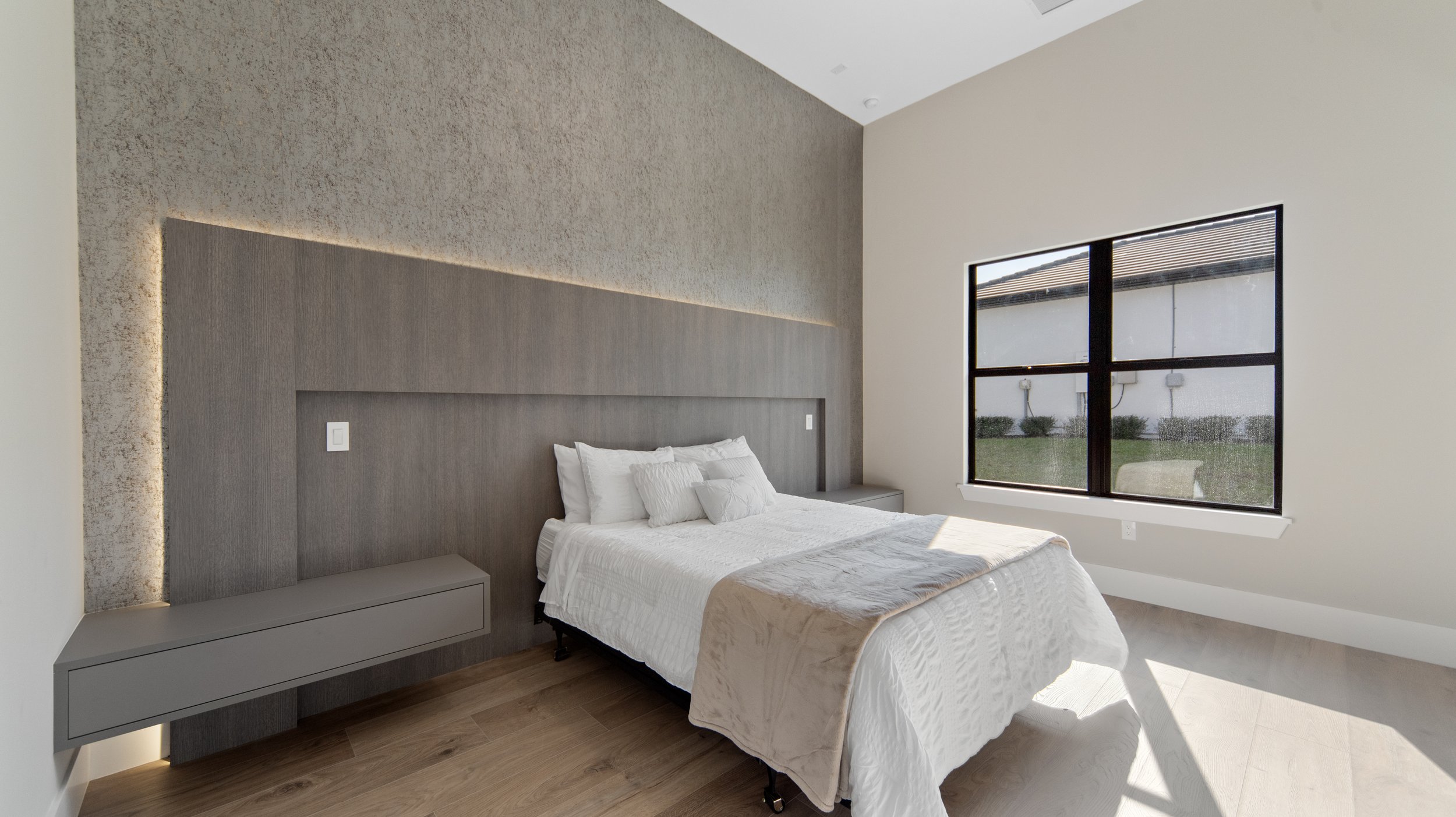 Minimalist bedroom with a bed, white bedding, a gray headboard and floating nightstands, large window with black frame, wood flooring, and neutral-colored walls.