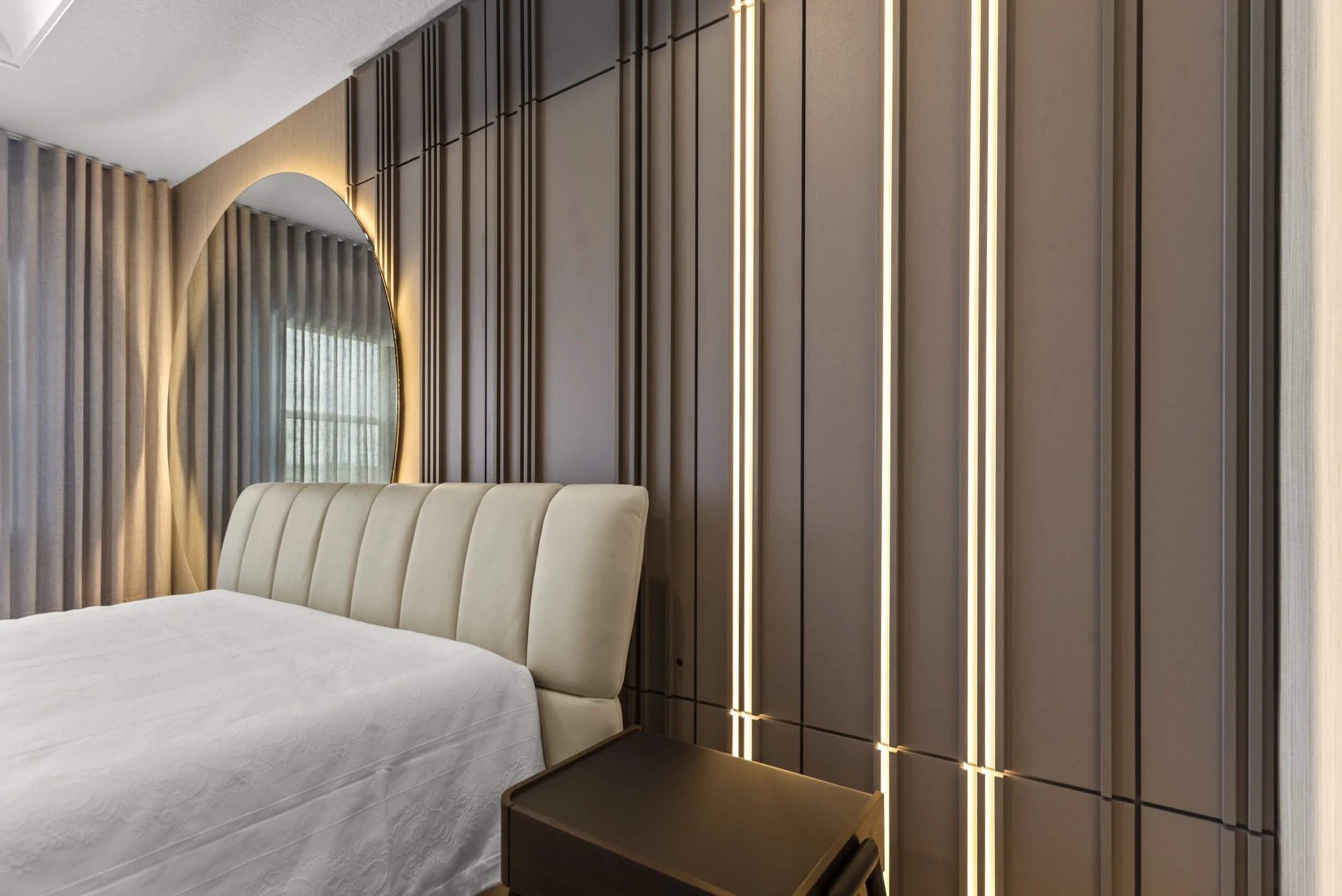 Modern bedroom with a cream upholstered headboard, geometric wall paneling with vertical LED lighting, and a brown nightstand.