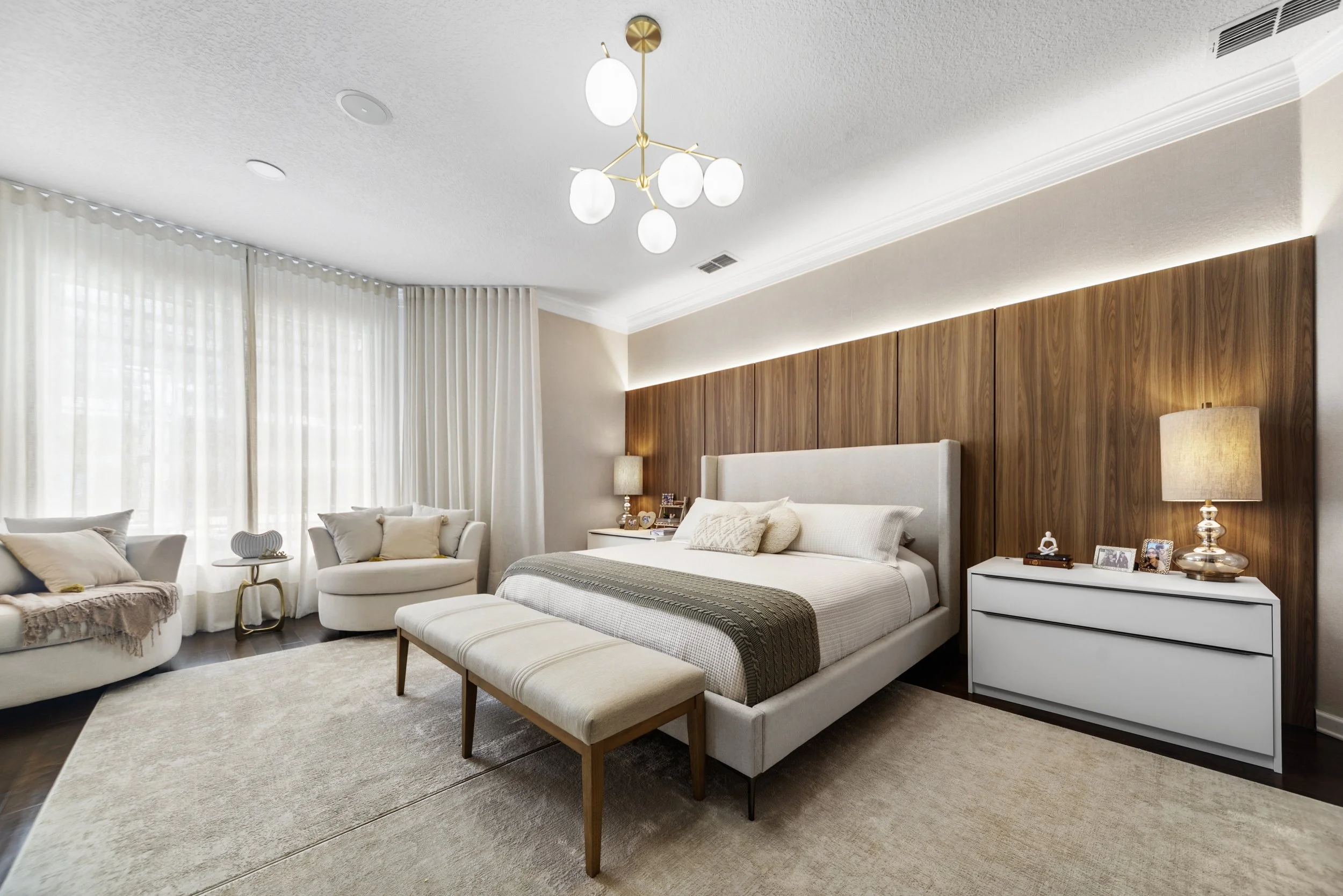 Modern bedroom with a large bed, wooden accent wall, bedside tables with lamps, a bench at the foot of the bed, and a sitting area with chairs and a small table near large windows with white curtains.