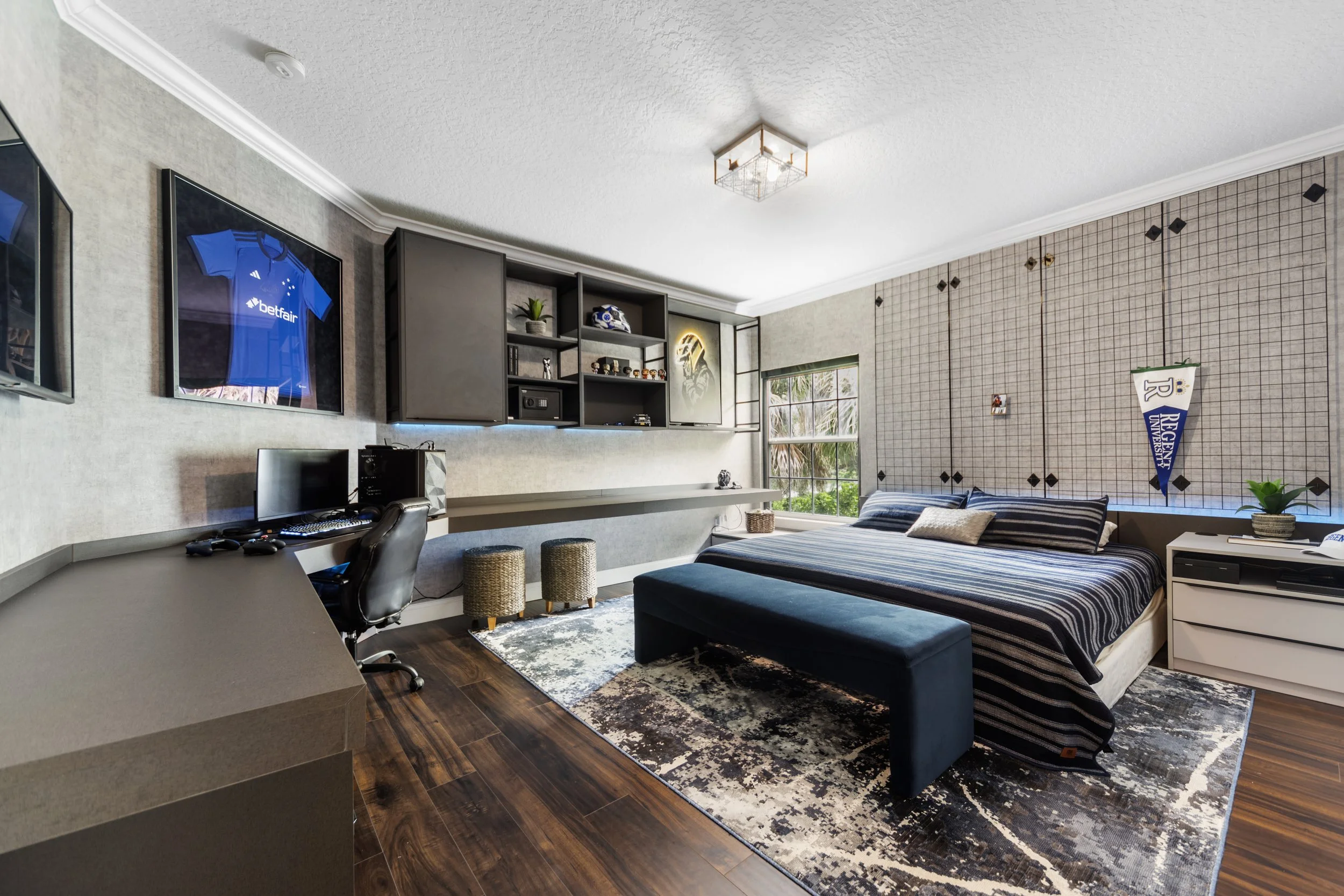 A bedroom with a bed, desk, computer, wall-mounted cabinets, window, and sports memorabilia.