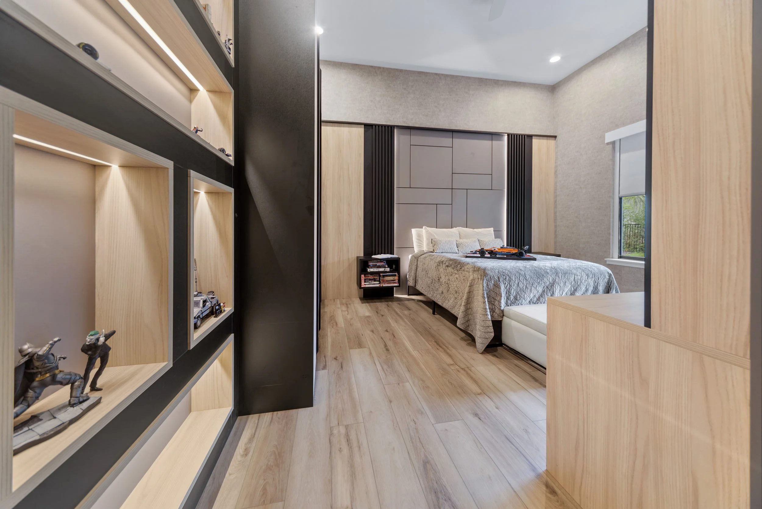 A modern bedroom with a bed, side tables, and a window. Wooden and grey wall panels decorate the headboard area, with toy figures on the shelf in the foreground.