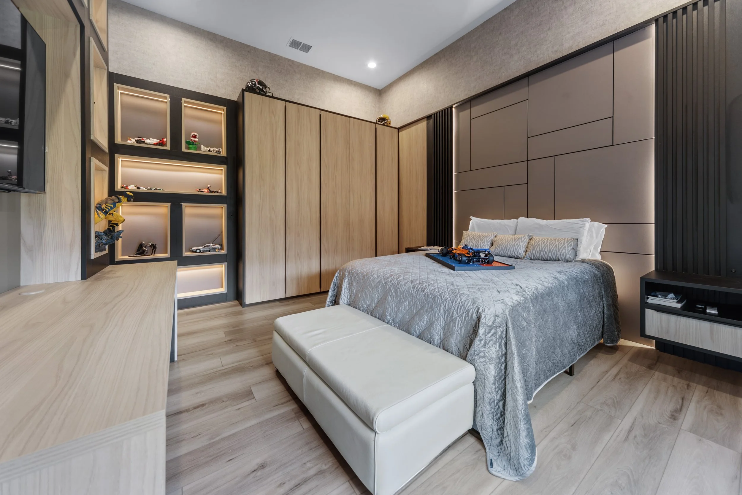 Modern bedroom with a bed, pillows, a bench at the foot of the bed, wood and dark paneled wall accents, light wood flooring, and built-in shelves with display car models.
