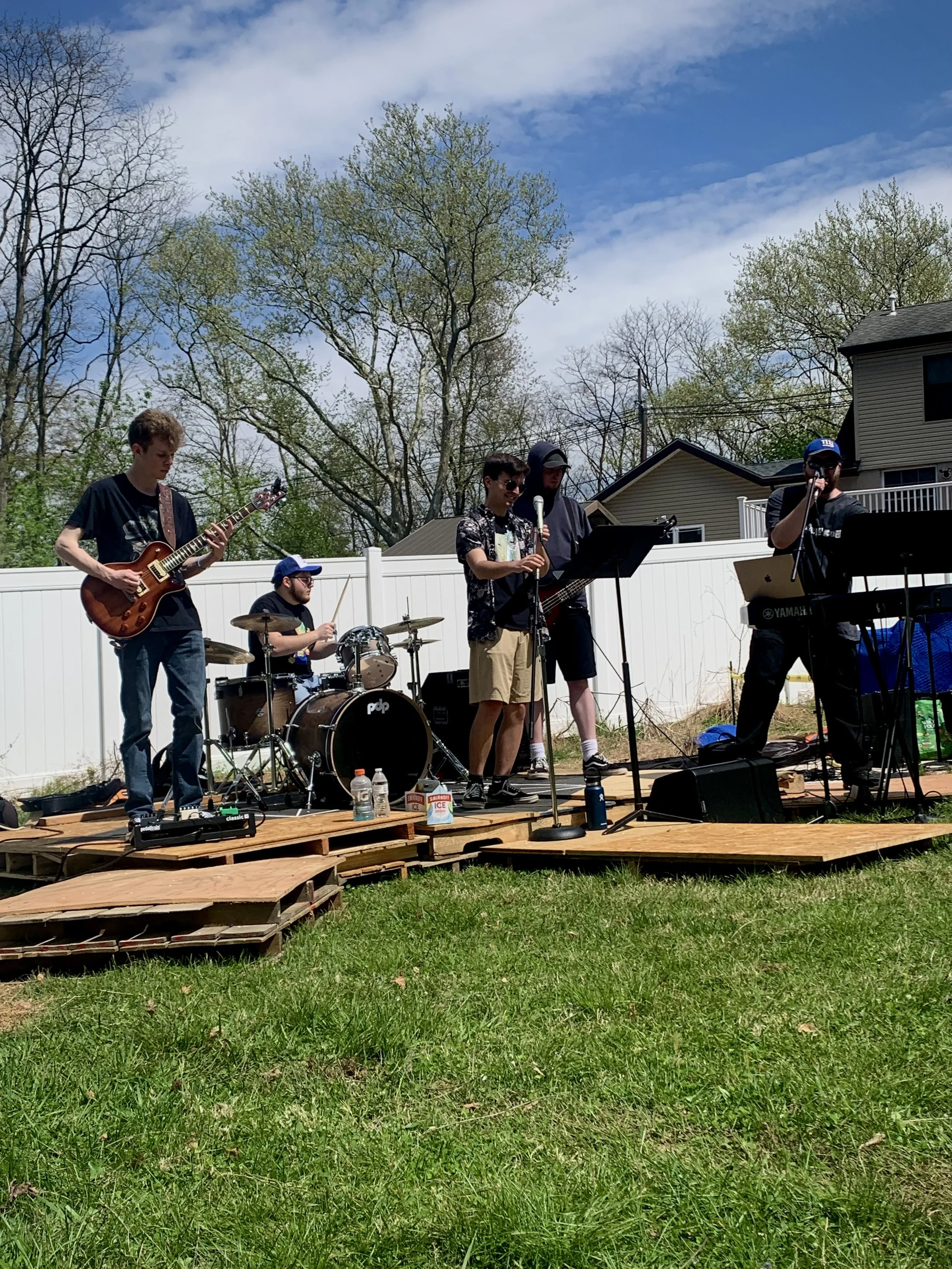 A group of five young men performing a band on a small wooden stage outdoors on a sunny day with a blue sky and trees in the background.