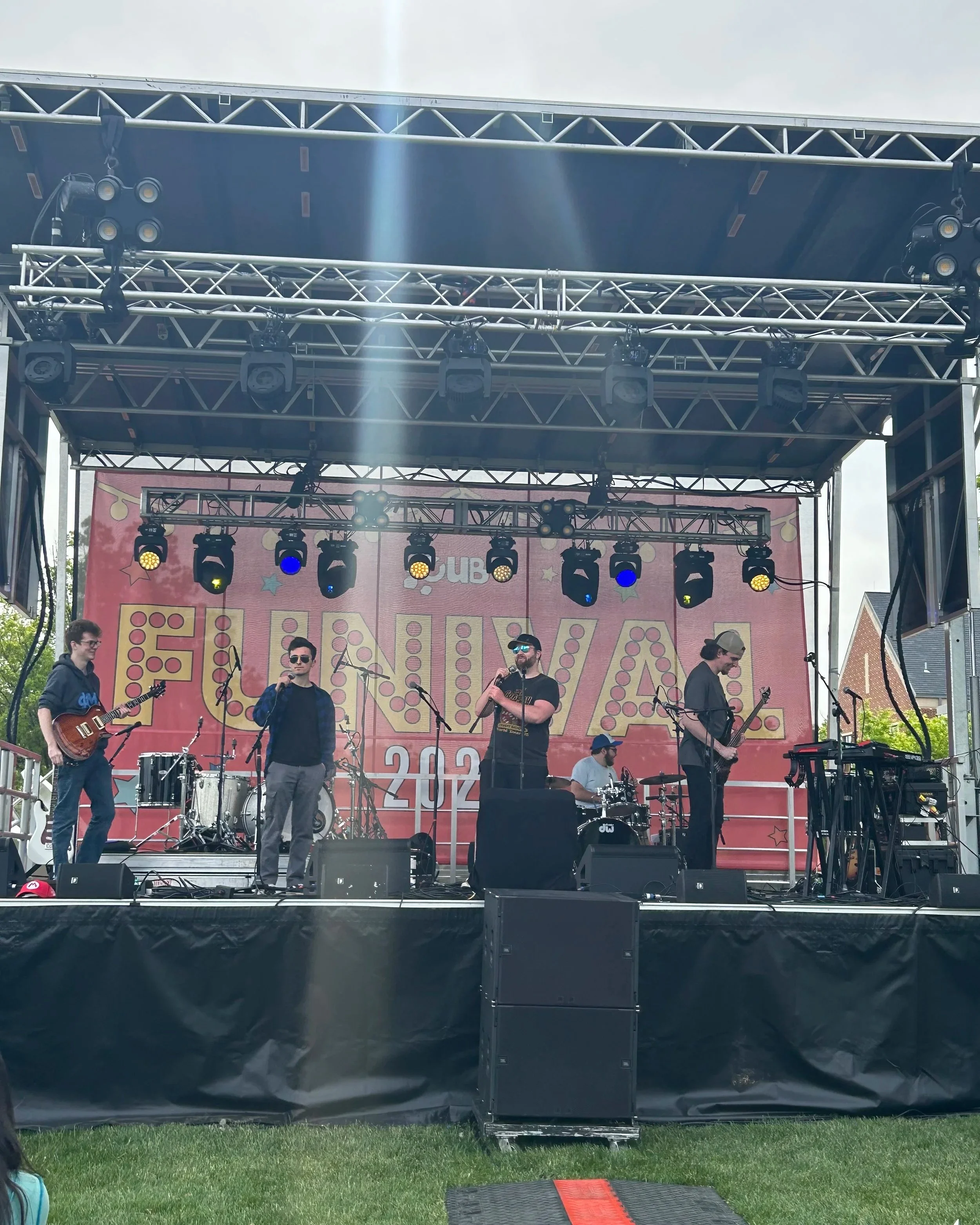 A live band performing on an outdoor stage with musicians playing various instruments and a colorful sign in the background.