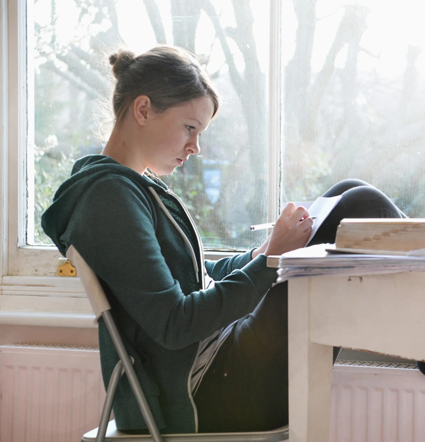 A young woman sitting on a chair by a window, reading a notebook