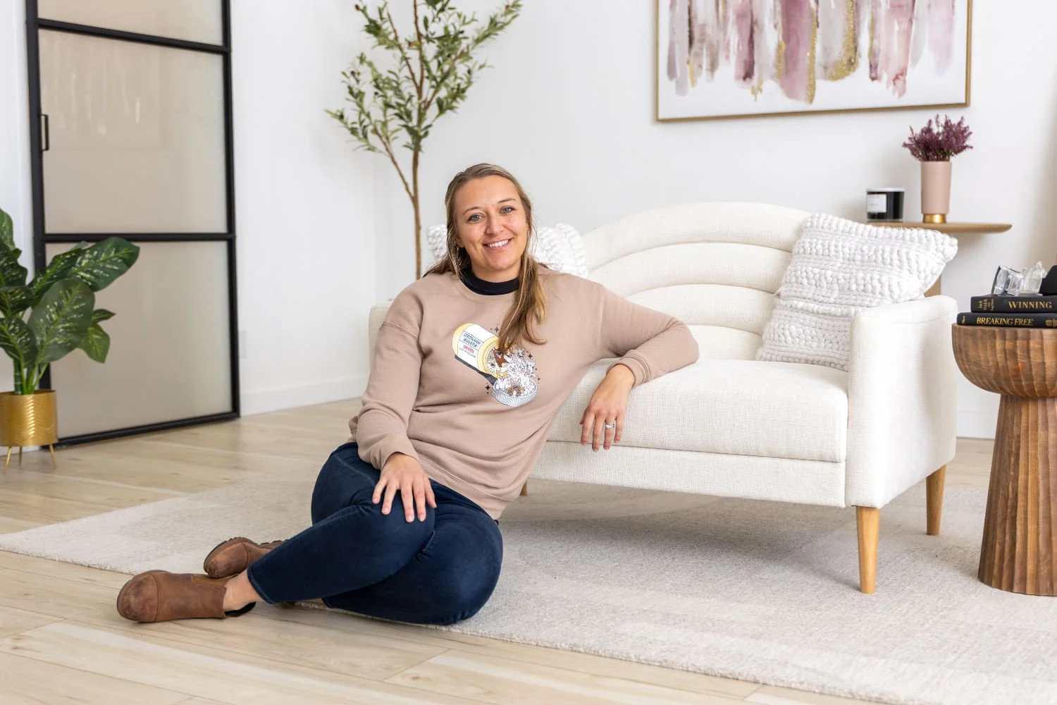 Ashley Braund sitting on the floor next to a white sofa in a modern living room, smiling at the camera.