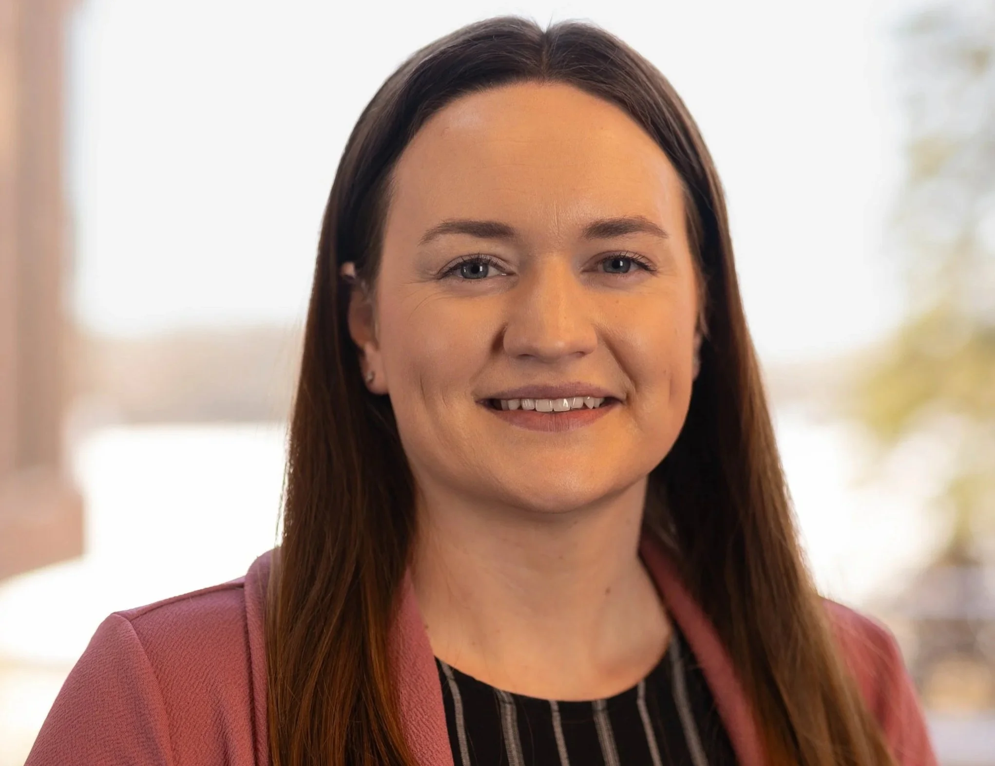 A young woman with long brown hair smiling, wearing a white top, in front of a wooden background.