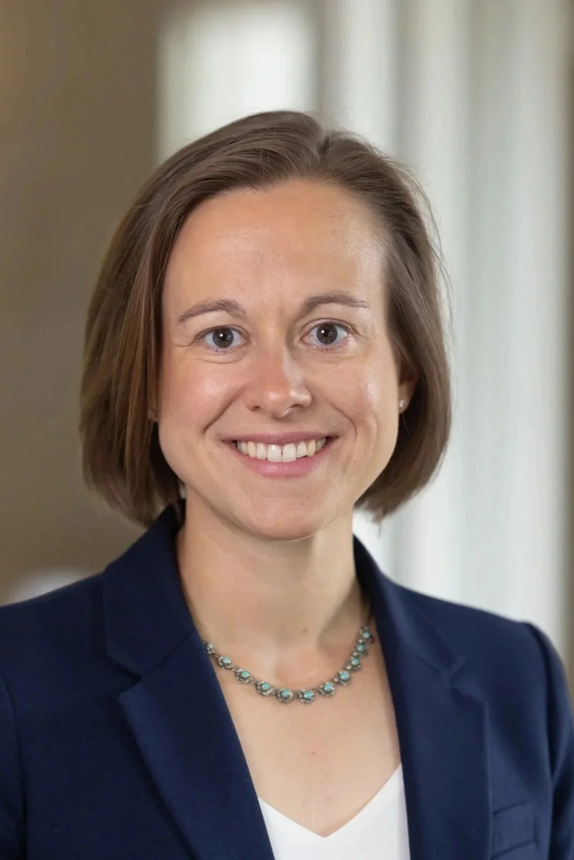 Woman with short brown hair, glasses, smiling, wearing a purple cardigan and gray top, standing against a wooden background.