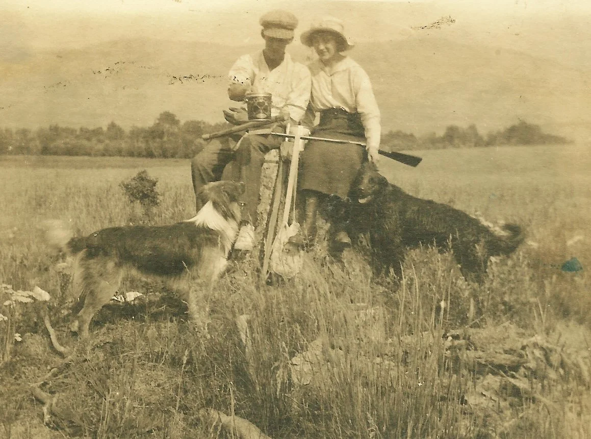 sepia toned photograph of young couple eating lunch together on a large stump with two large dogs circa 1910.