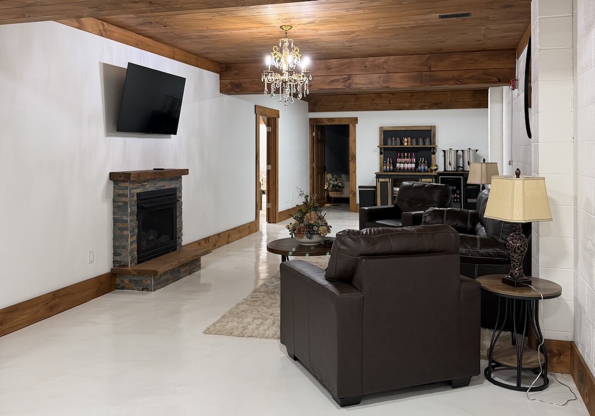Cozy living room with white walls, wooden ceiling and trim, black leather sofa, matching armchair, table lamp, floral centerpiece on a coffee table, wall-mounted TV, fireplace with brick and wood mantle, and a bar area with shelves and coffee urns in