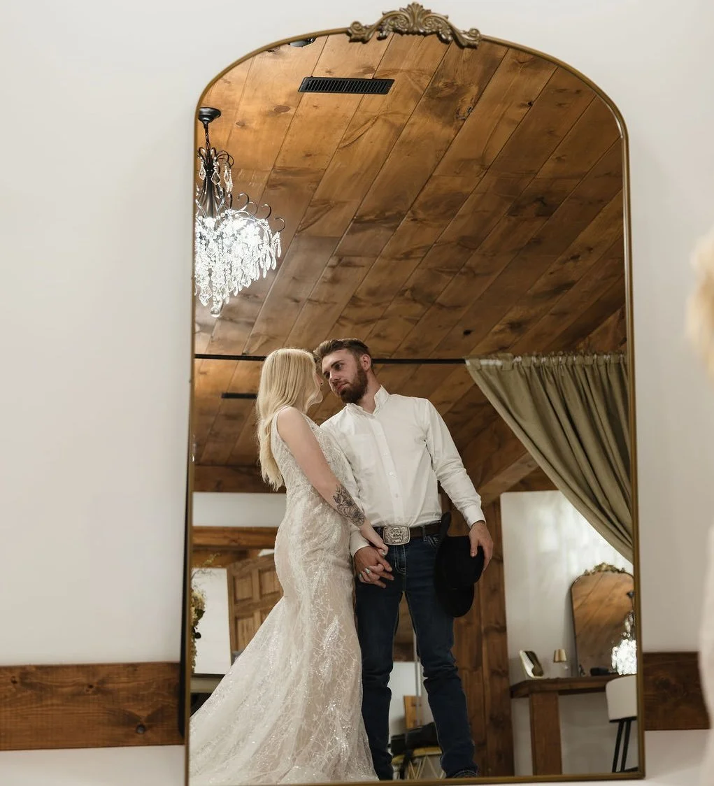 A bride and groom holding hands and looking at each other, reflected in a mirror with a wooden ceiling and chandelier above, during their wedding or engagement photos.