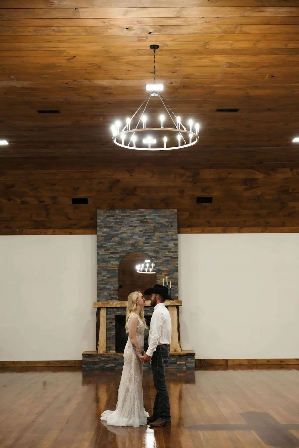 A bride and groom holding hands, facing each other in a rustic indoor setting with wooden ceiling and floor, stone fireplace, and chandelier.