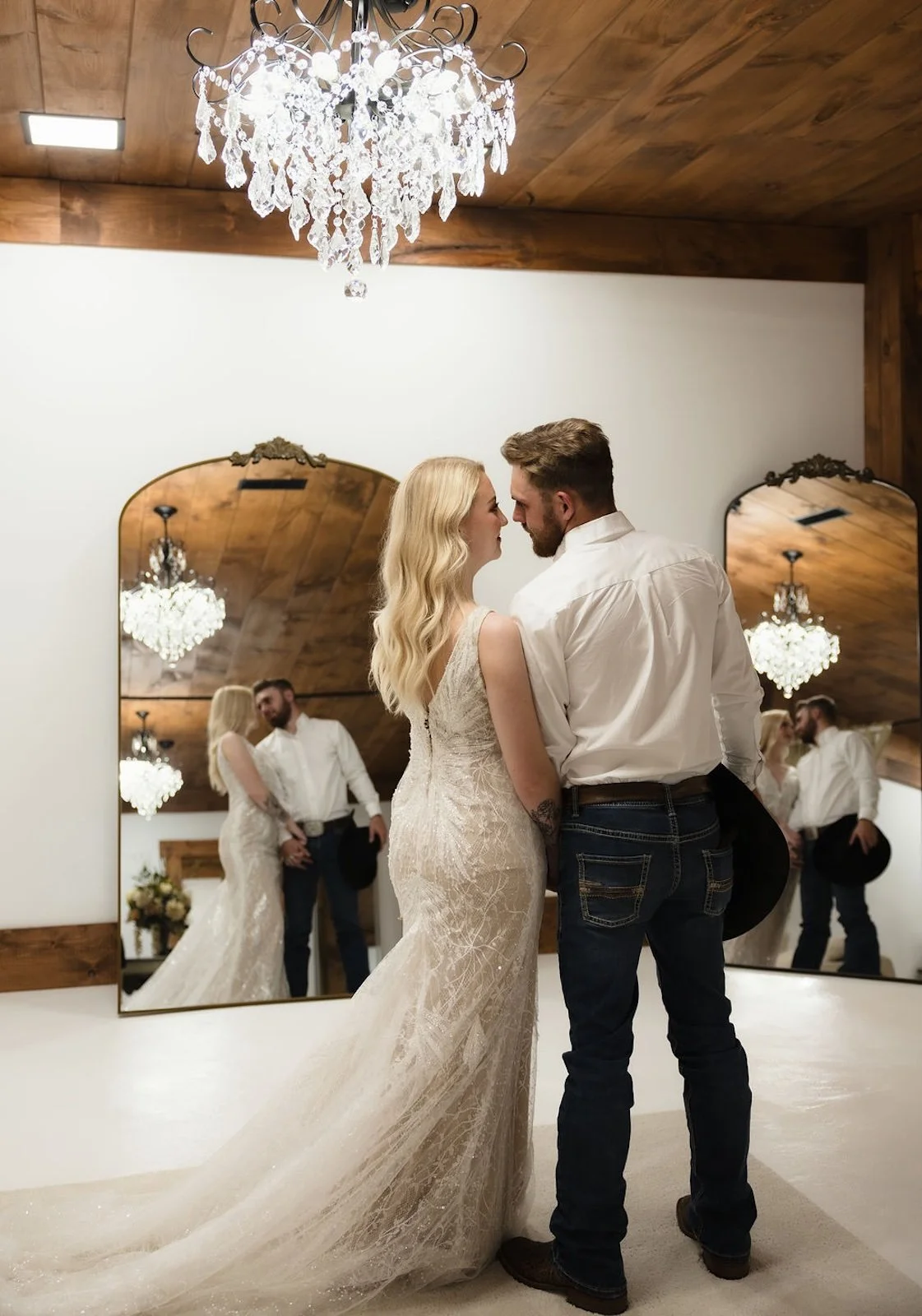 A bride and groom standing close, looking into each other's eyes, in front of a large mirror in a room with wooden ceiling and walls, a chandelier hanging above them, and the reflection of their intimate moment.