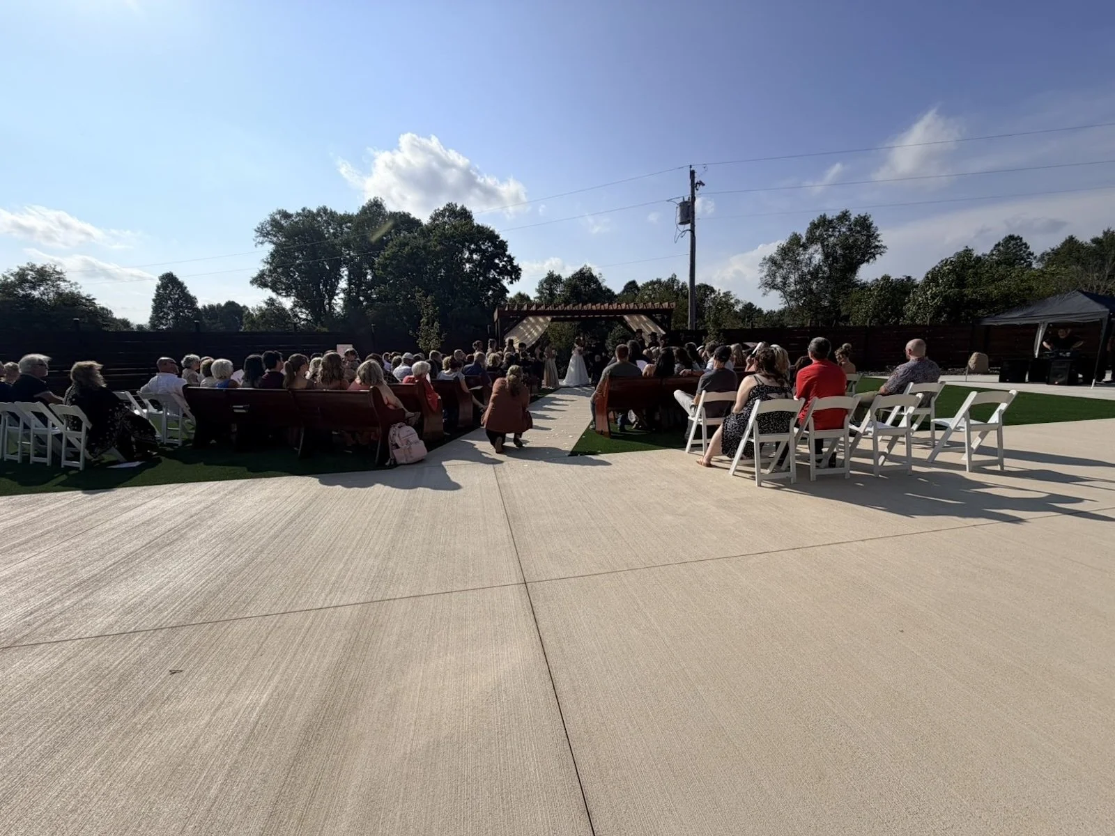 Outdoor wedding ceremony with guests seated on benches and chairs, officiant and bride visible at the altar under a wooden pergola, sunny day with blue sky and some clouds.