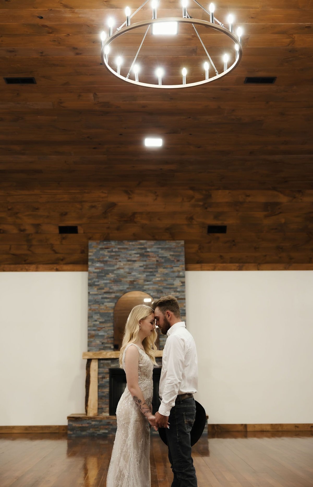 A couple standing close with foreheads touching in an indoor setting with wooden ceiling, chandelier, and brick fireplace