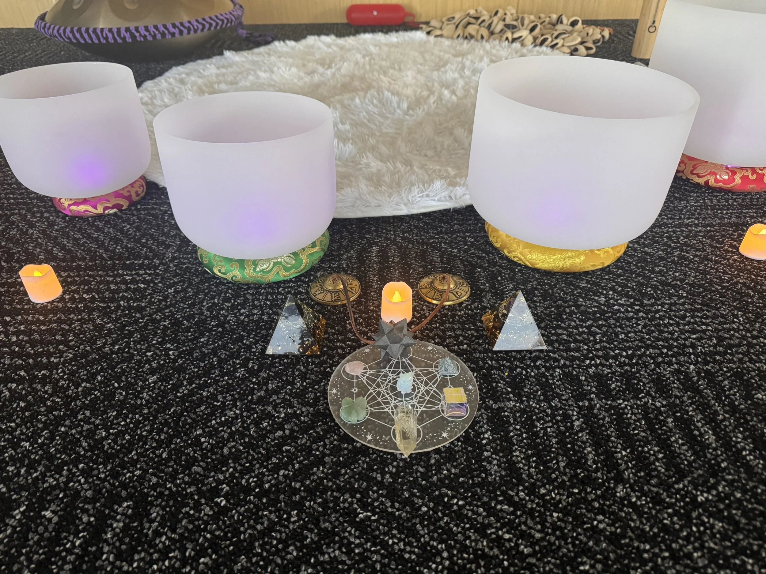 Sound bath setup with crystal singing bowls, tingsha bell, and handpan. Crystals laid out in sacred geometry with orgone pyramids surrounding it with electronic candles.